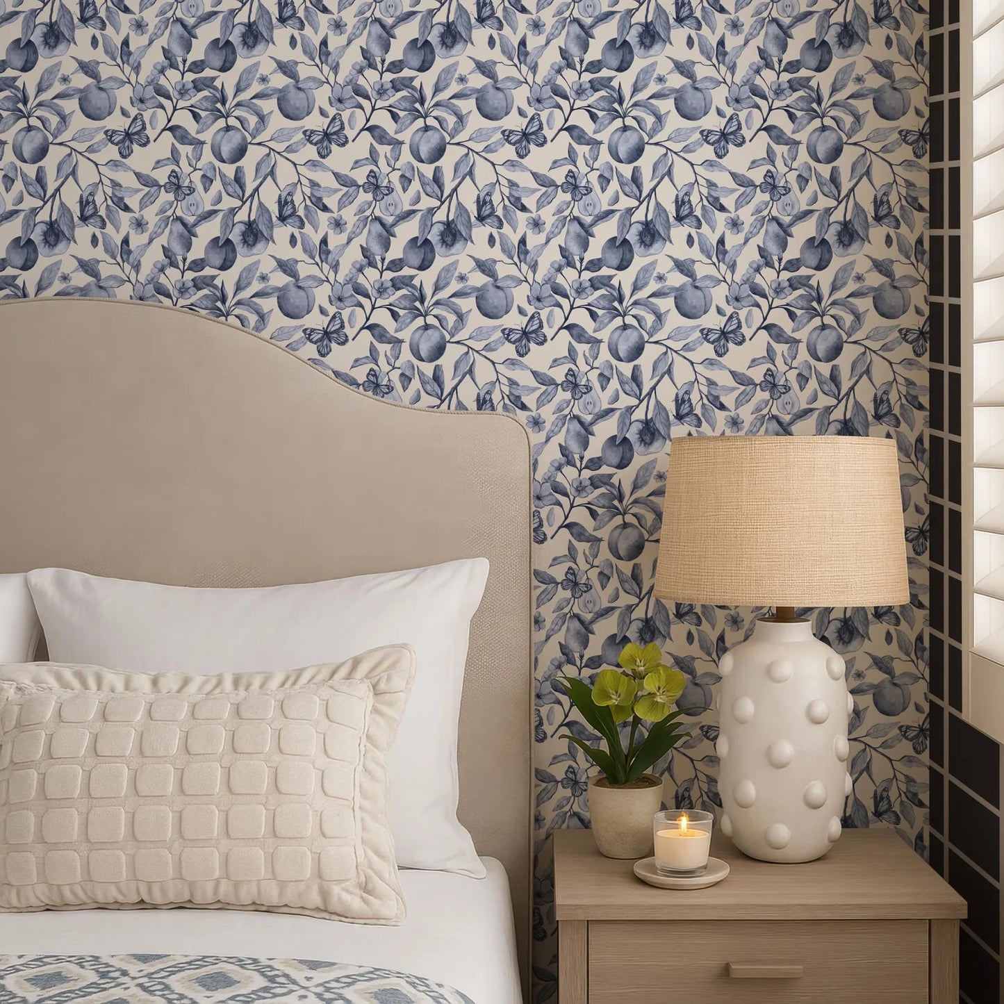 a cozy bedroom scene with a bed, a nightstand, and a lamp. The bed has a beige headboard and white pillows, and the nightstand has a white lamp with a beige shade. The wall behind the bed is covered in a blue and white floral wallpaper.