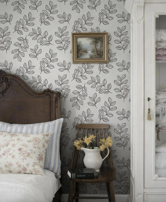 a bedroom with a wooden headboard, a white bedspread, and a wooden chair with a vase of yellow flowers. The wall behind the chair has a black and white leaf pattern wallpaper, and there is a framed picture hanging above the chair.