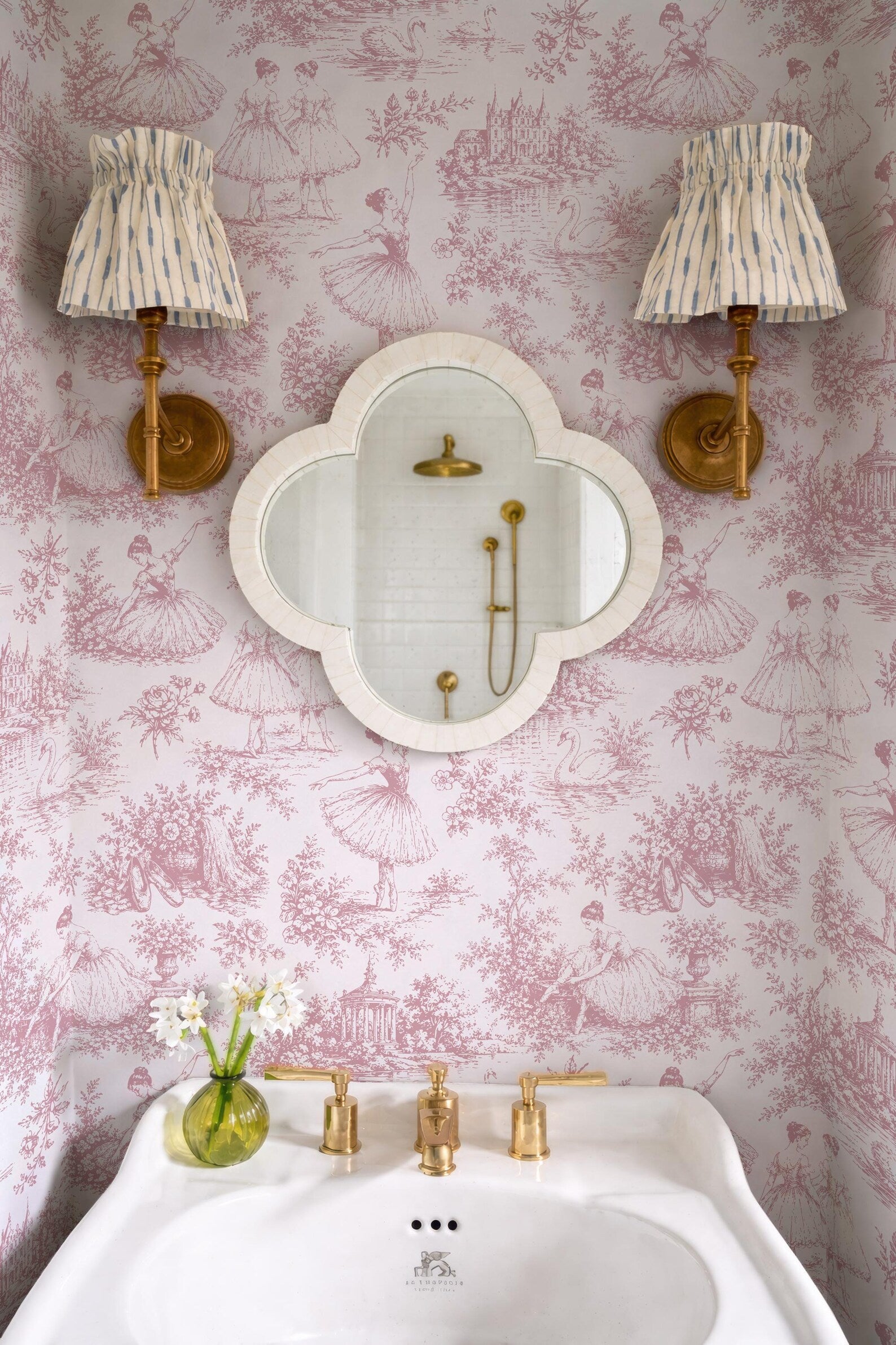 a bathroom with pink and white wallpaper, a white sink with gold fixtures, and two wall sconces with striped fabric shades. There is also a mirror above the sink.