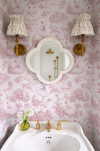a bathroom with pink and white wallpaper, a white sink with gold fixtures, and two wall sconces with striped fabric shades. There is also a mirror above the sink.