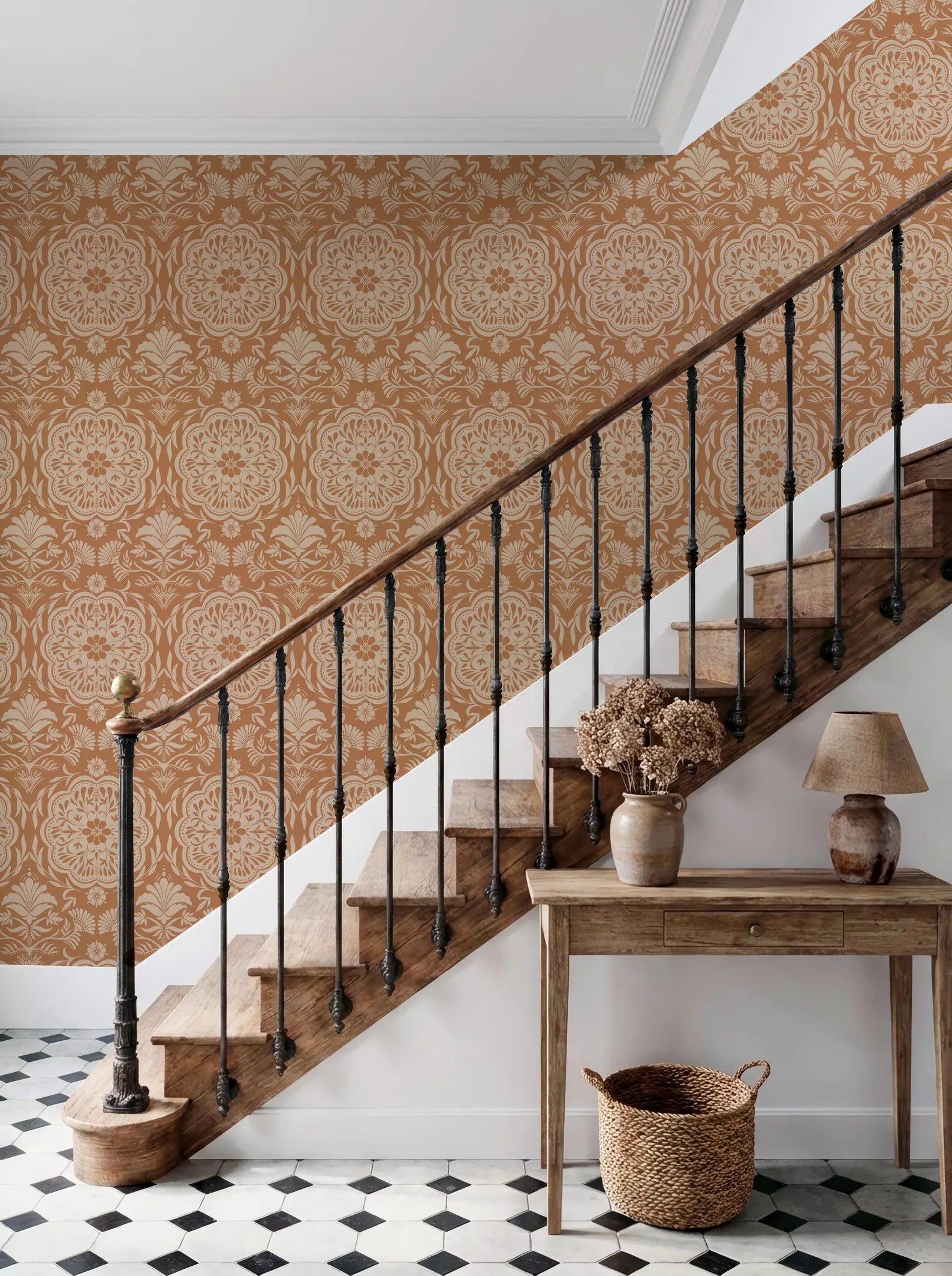 a wooden staircase with a black metal railing, set against a wall with a repeating pattern of orange and white damask wallpaper. The staircase leads up to a wooden table with a lamp and a vase of flowers, situated on a black and white checkered floor.