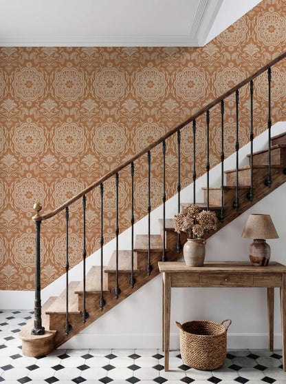 a wooden staircase with a black metal railing, set against a wall with a repeating pattern of orange and white damask wallpaper. The staircase leads up to a wooden table with a lamp and a vase of flowers, situated on a black and white checkered floor.