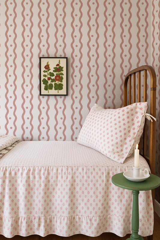 a cozy bedroom scene with a bed, a wooden headboard, and a framed floral artwork on the wall. The bed is covered in a white and pink patterned bedspread, and there is a green side table with a white candle on it. The overall color scheme and arrangement of the elements create a warm and inviting atmosphere.
