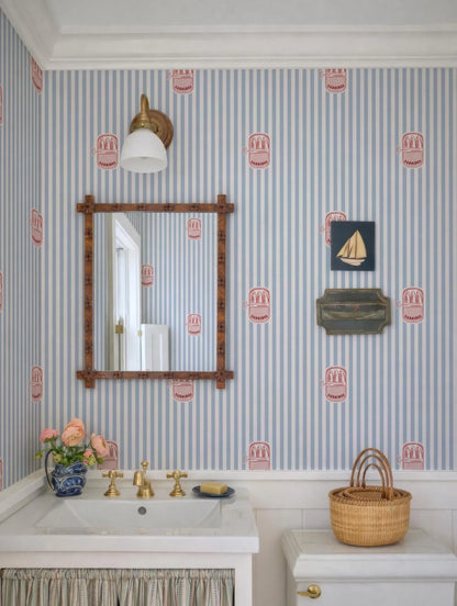 a bathroom with a white sink, a mirror, and a wooden frame. The walls are covered in blue and white striped wallpaper with red and white designs. There is a wicker basket on the toilet tank and a vase of flowers on the sink.