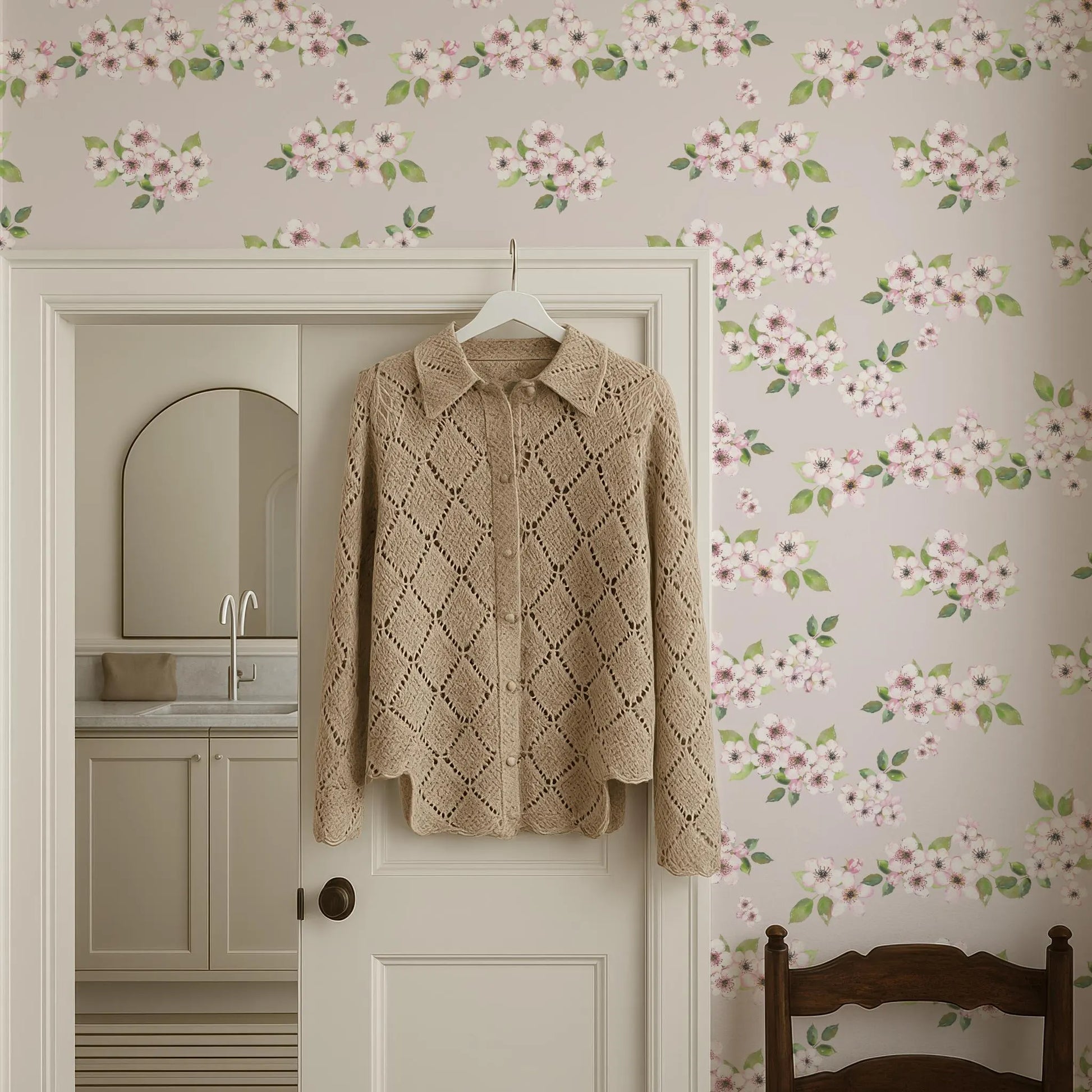 a cozy, beige knitted sweater hanging on a white door, with a floral wallpaper behind it.