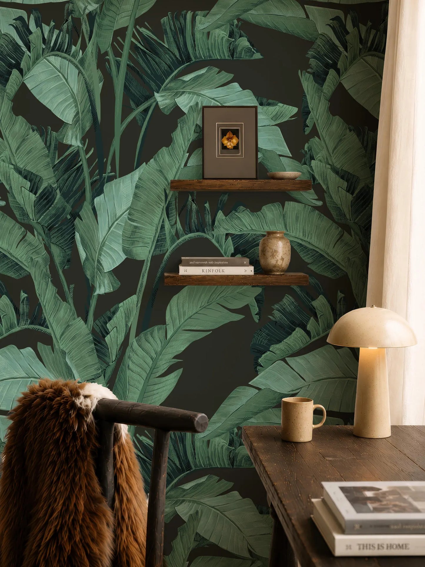 a cozy and stylish living space with a large wallpapered wall that has a tropical leaf pattern. The room includes a wooden desk with a lamp, a shelf with books and a vase, and a brown fur coat draped over a chair.