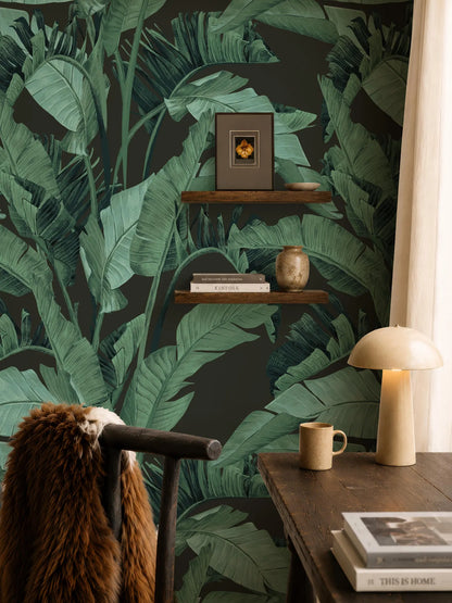 a cozy and stylish living space with a large wallpapered wall that has a tropical leaf pattern. The room includes a wooden desk with a lamp, a shelf with books and a vase, and a brown fur coat draped over a chair.