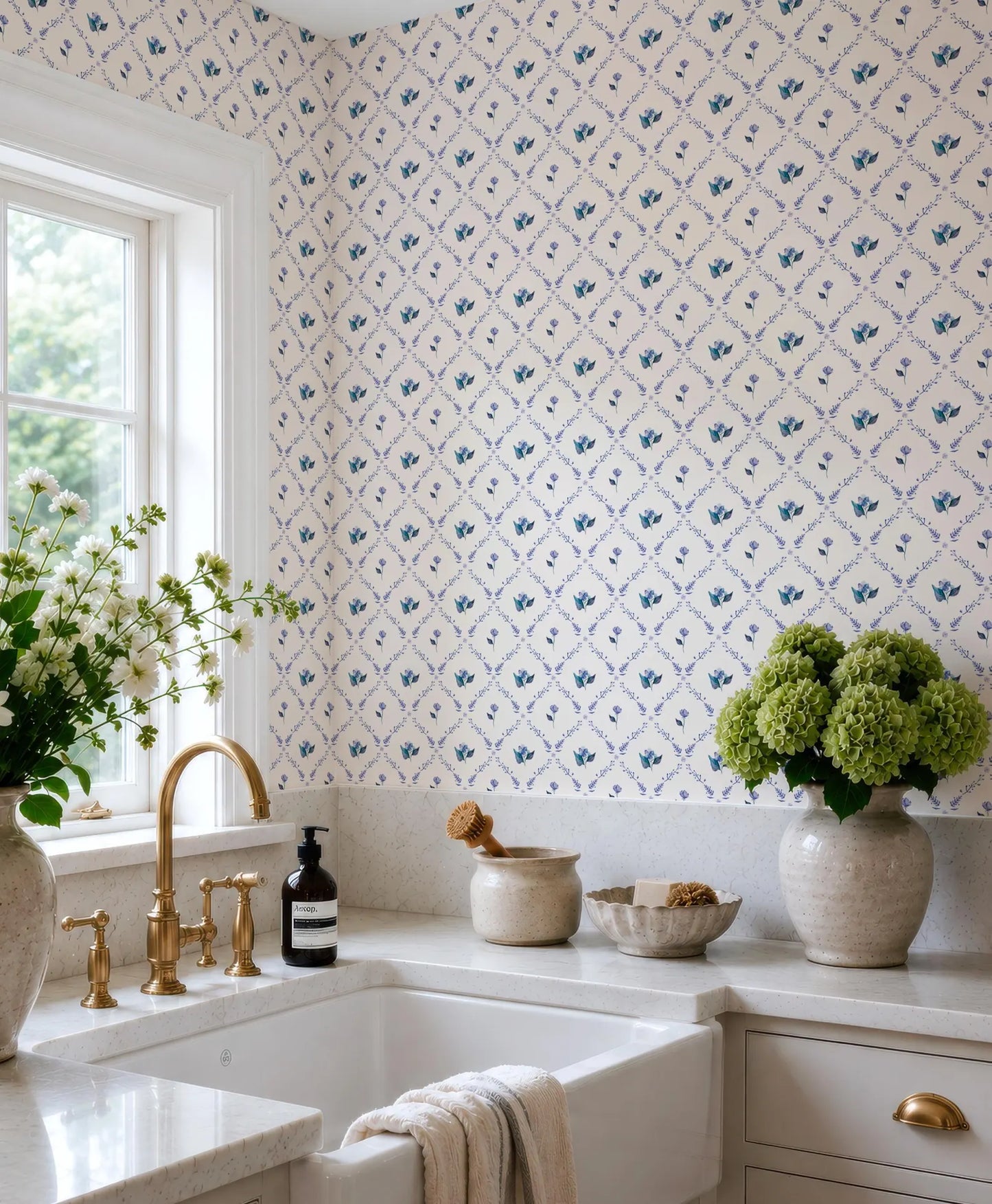 a well-organized kitchen with a white sink, a vase of green hydrangeas, and a bottle of soap on the counter. The walls are adorned with a blue and white floral wallpaper, and there is a window above the sink.