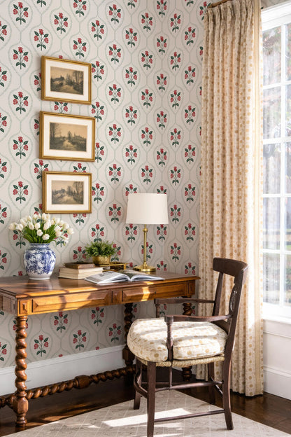 a cozy corner of a room with a wooden desk, a lamp, and a chair. The wall behind the desk is decorated with a floral wallpaper pattern. There are three framed pictures hanging on the wall, and a vase of flowers on the desk. The room has a window with curtains, and the overall atmosphere appears warm and inviting.