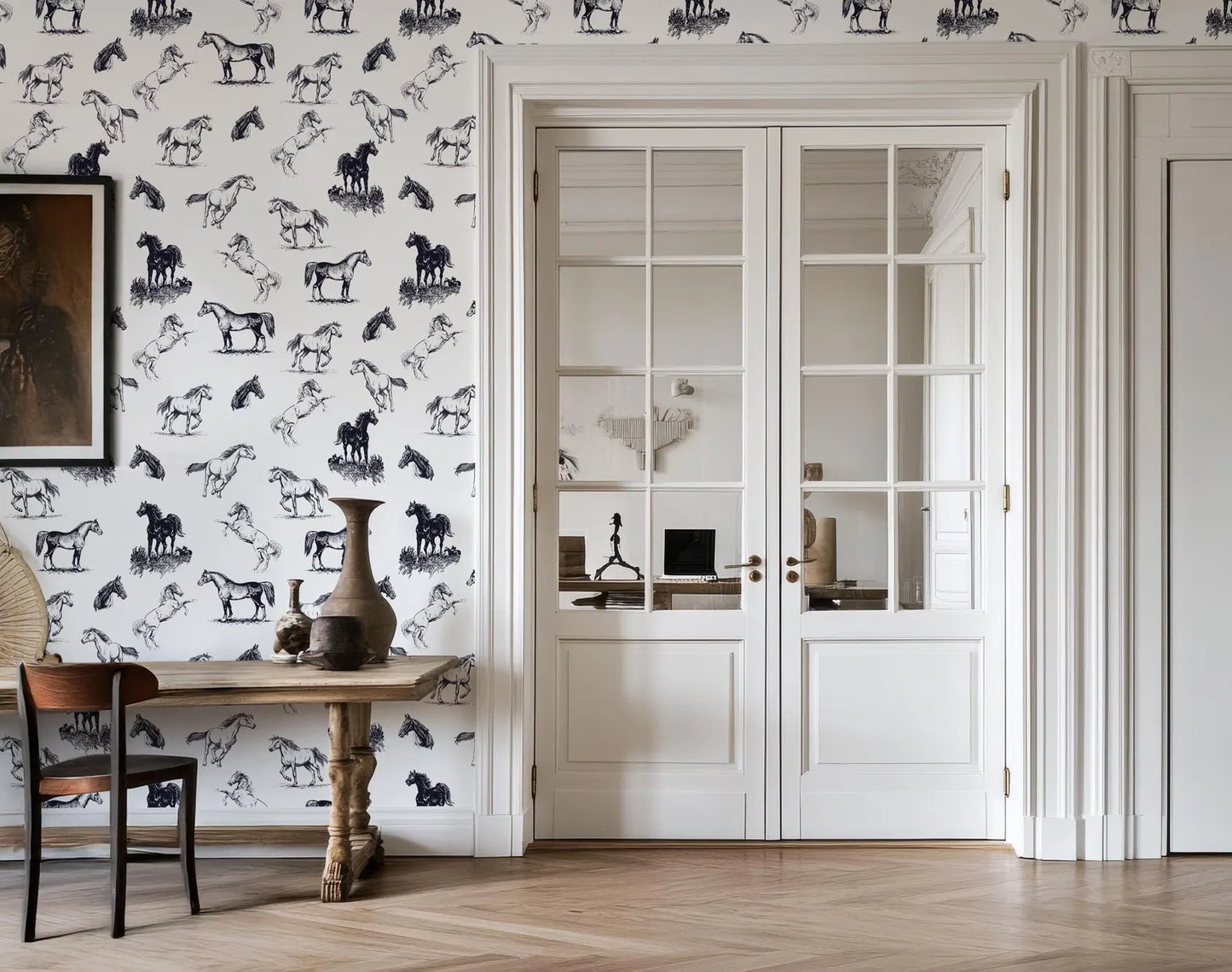 a room with a white door and a wooden table with a vase on it. The wall behind the table has a pattern of black and white horses. There is also a chair in the room.