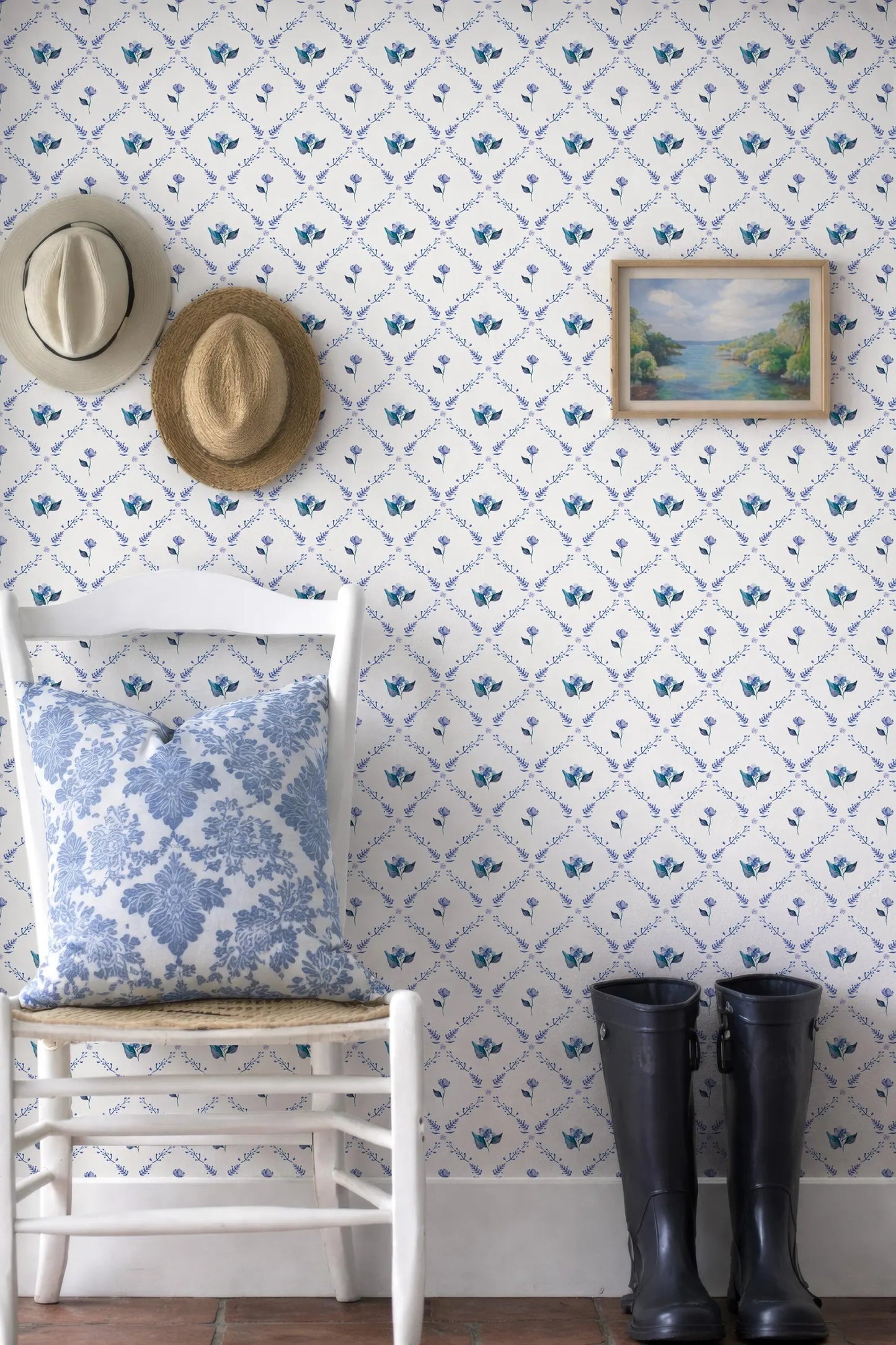 a cozy corner with a white chair, a pair of black rain boots, and a framed picture of a landscape on the wall. The chair has a blue and white floral pillow on it.