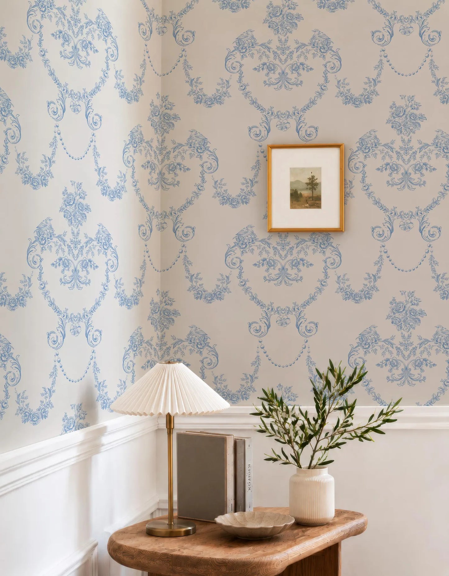 a corner of a room with a wooden table, a lamp, a framed picture, and a vase with greenery. The wallpaper has a blue floral pattern, and the table is set against a white wall.