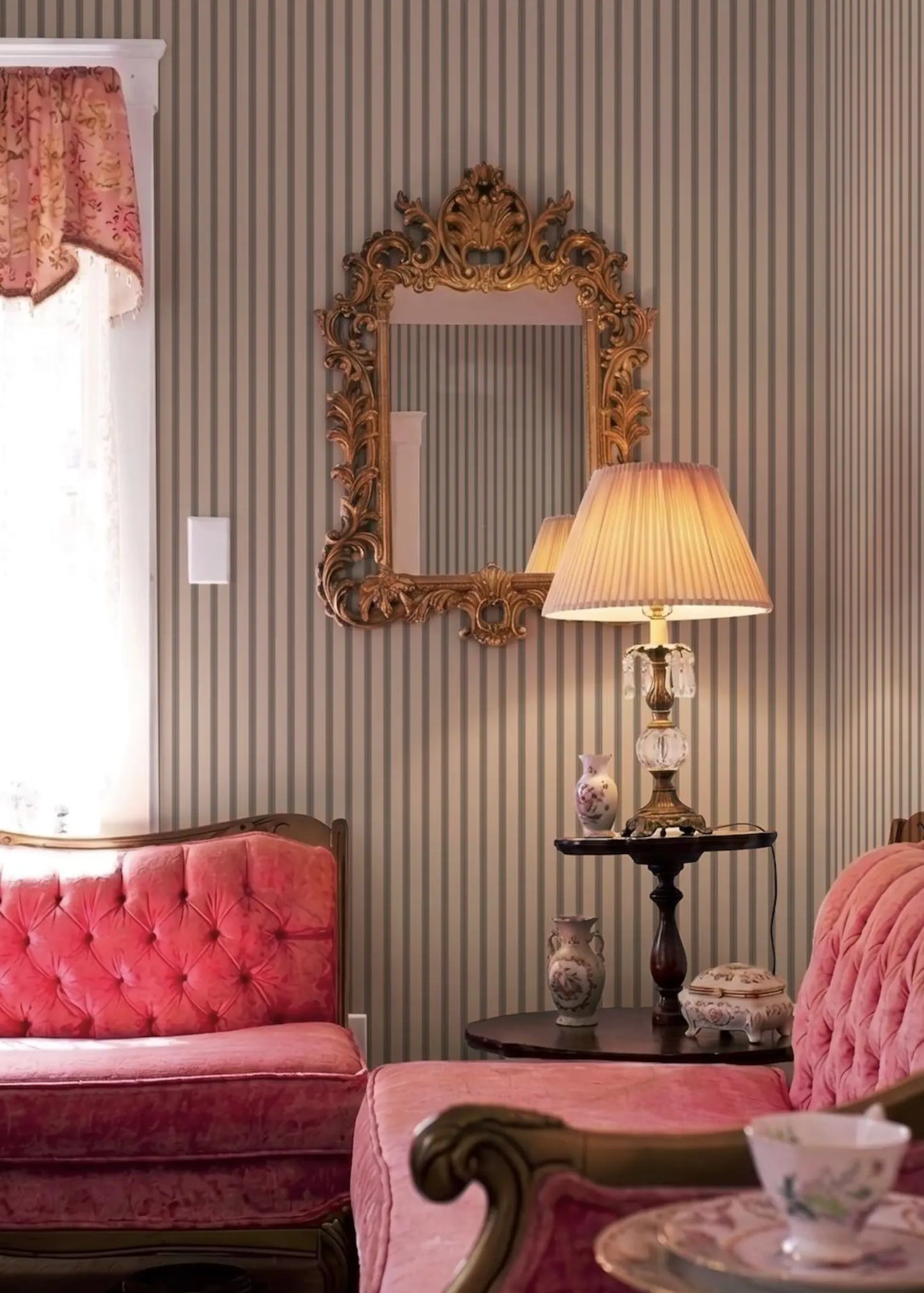 a cozy and elegant living room with a pink tufted sofa, a wooden side table with a lamp and vases, and a large ornate mirror on the wall.