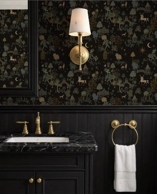 a bathroom with a black vanity, a gold faucet, and a black and gold wallpaper with a forest scene. There is a white towel hanging on a gold towel ring, and a gold wall sconce is mounted above the sink.