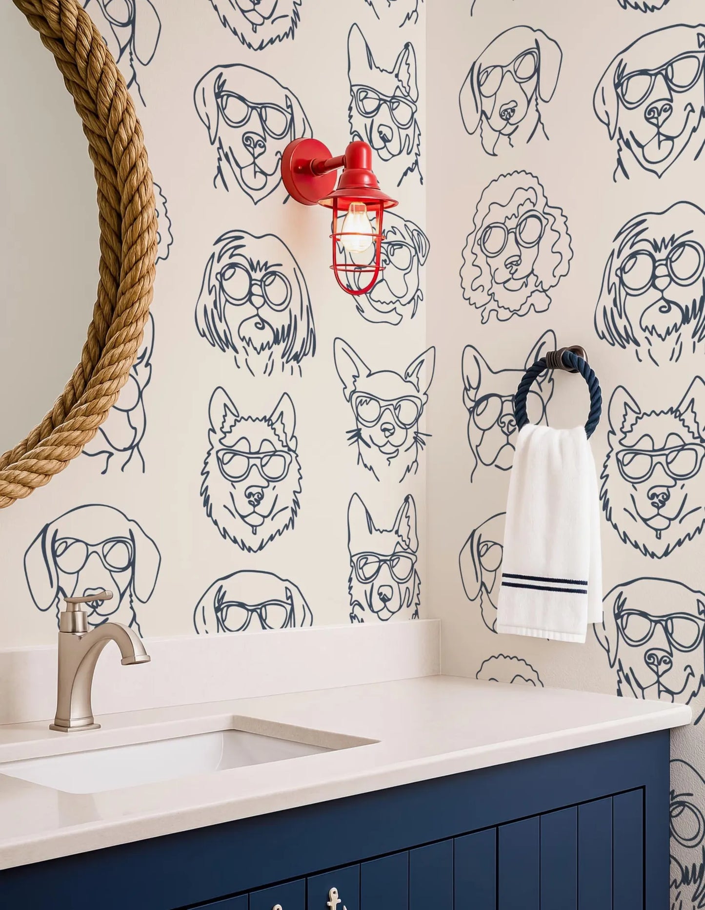a bathroom with a blue vanity and a wallpapered wall with various dog illustrations. There is a red light fixture mounted on the wall, and a towel rack with a white towel hanging on it.