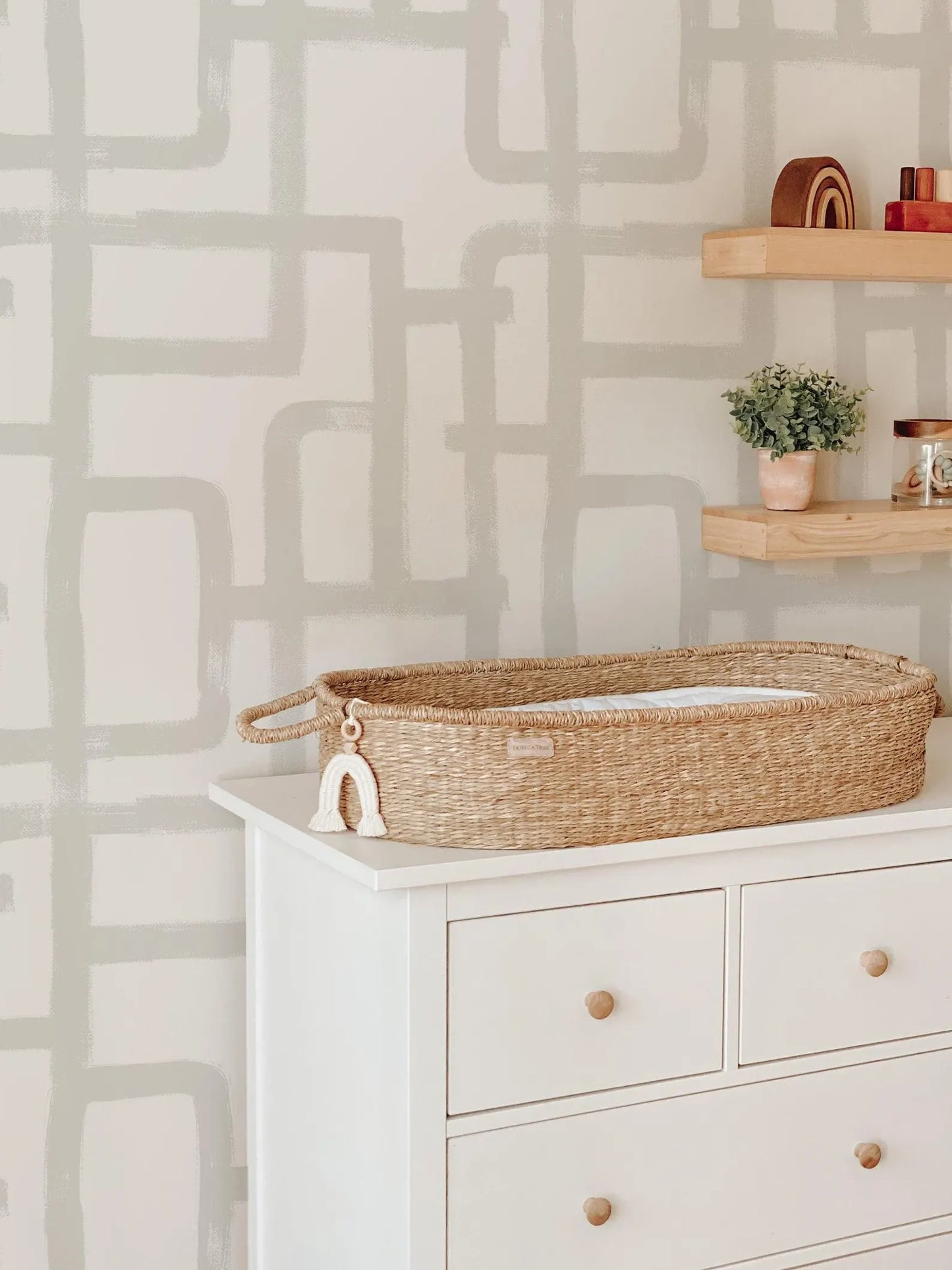 a white dresser with a woven basket on top, a wooden shelf above it, and a plant on the shelf. The dresser has three drawers and a wooden handle on the front. The background has a gray and white abstract pattern.