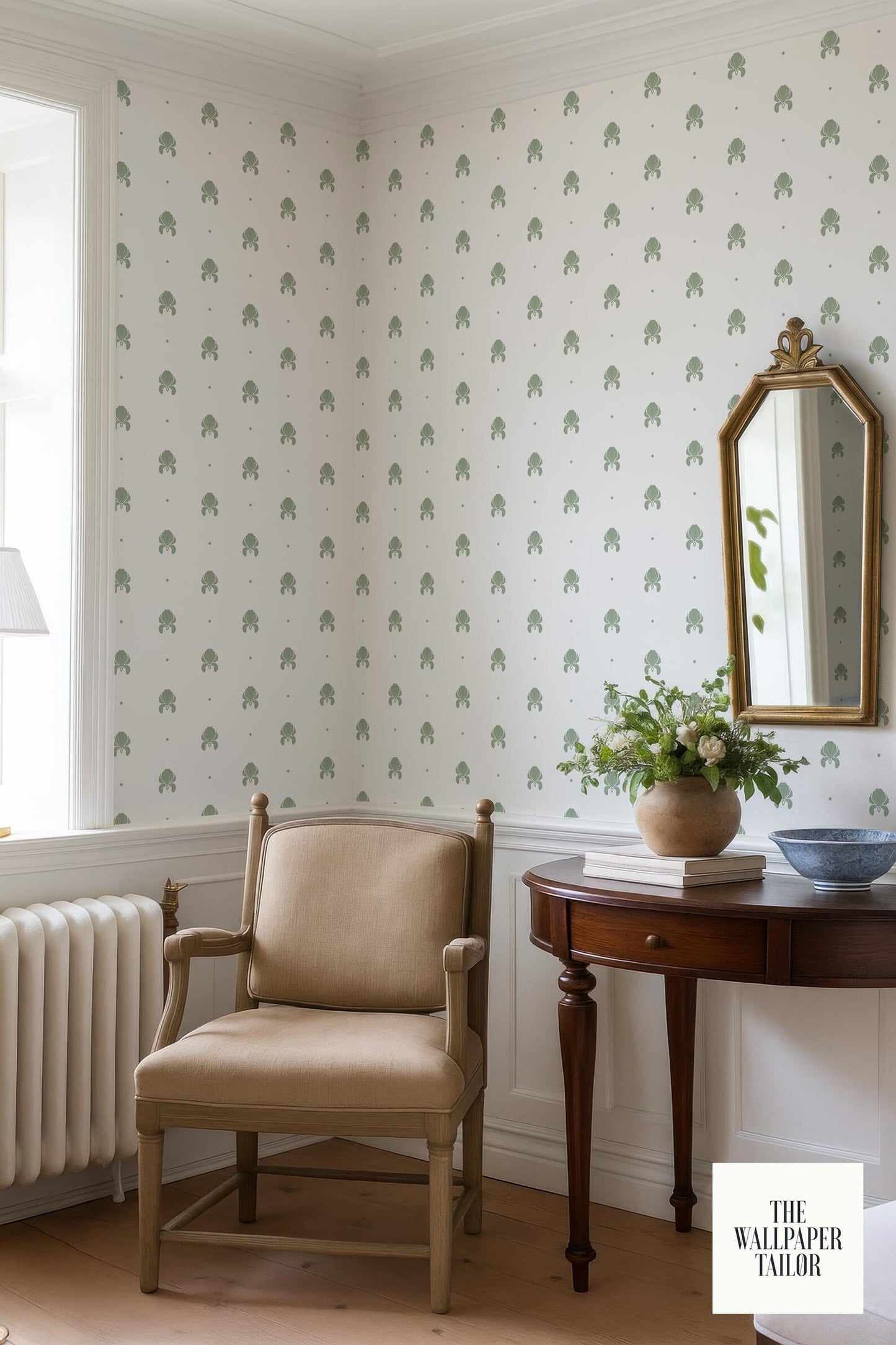 a cozy corner of a room with a beige armchair, a wooden side table with a vase of flowers, and a mirror above the table. The walls are covered in a patterned wallpaper with green trees, and there is a window in the background.