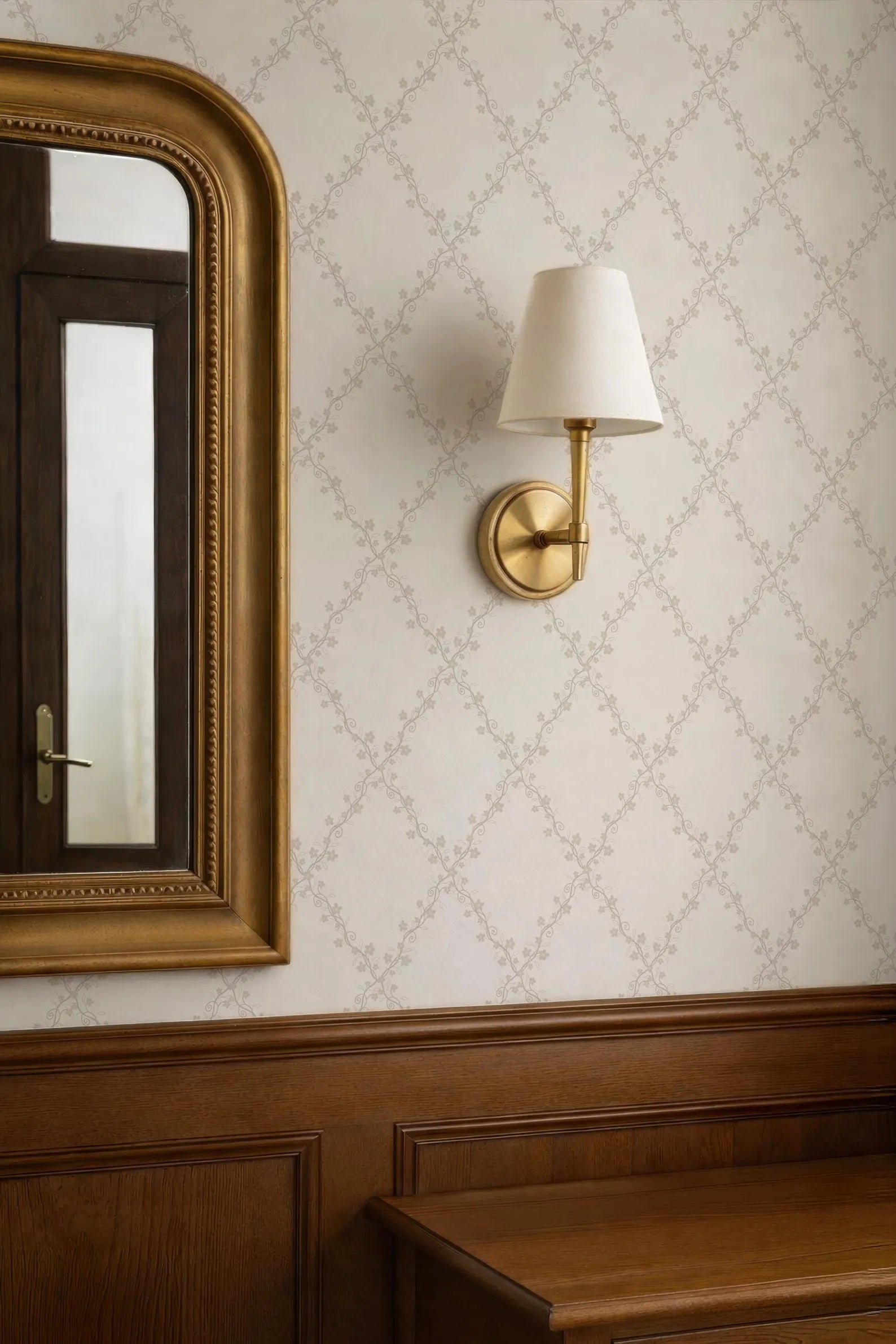 a wall with a gold-framed mirror and a gold-framed wall lamp, both mounted on a wall with a floral patterned wallpaper.