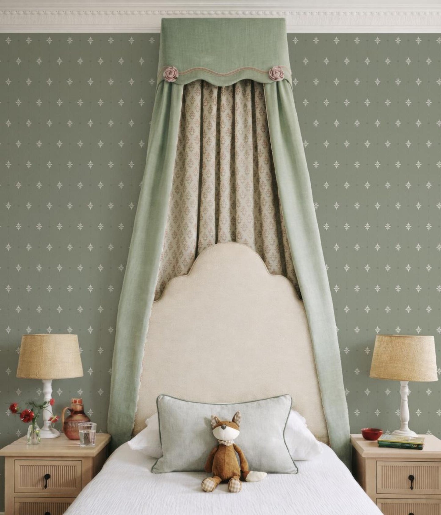 a cozy bedroom with a white bed, a nightstand on each side, and a canopy over the bed. The walls are covered in a patterned wallpaper, and there are two lamps on the nightstands. A stuffed animal, likely a teddy bear, is sitting on the bed.