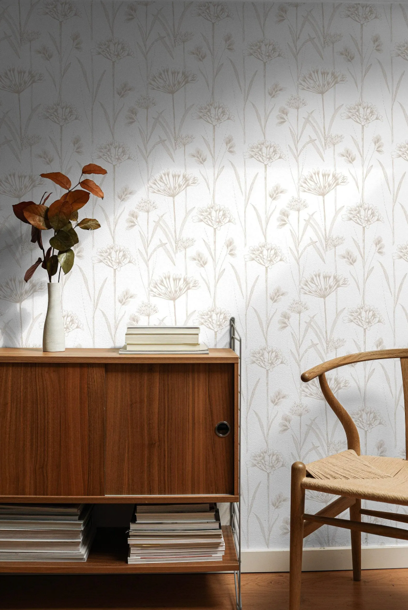 a wooden cabinet with a vase of dried flowers on top, a stack of books, and a woven chair in a room with a floral wallpaper.
