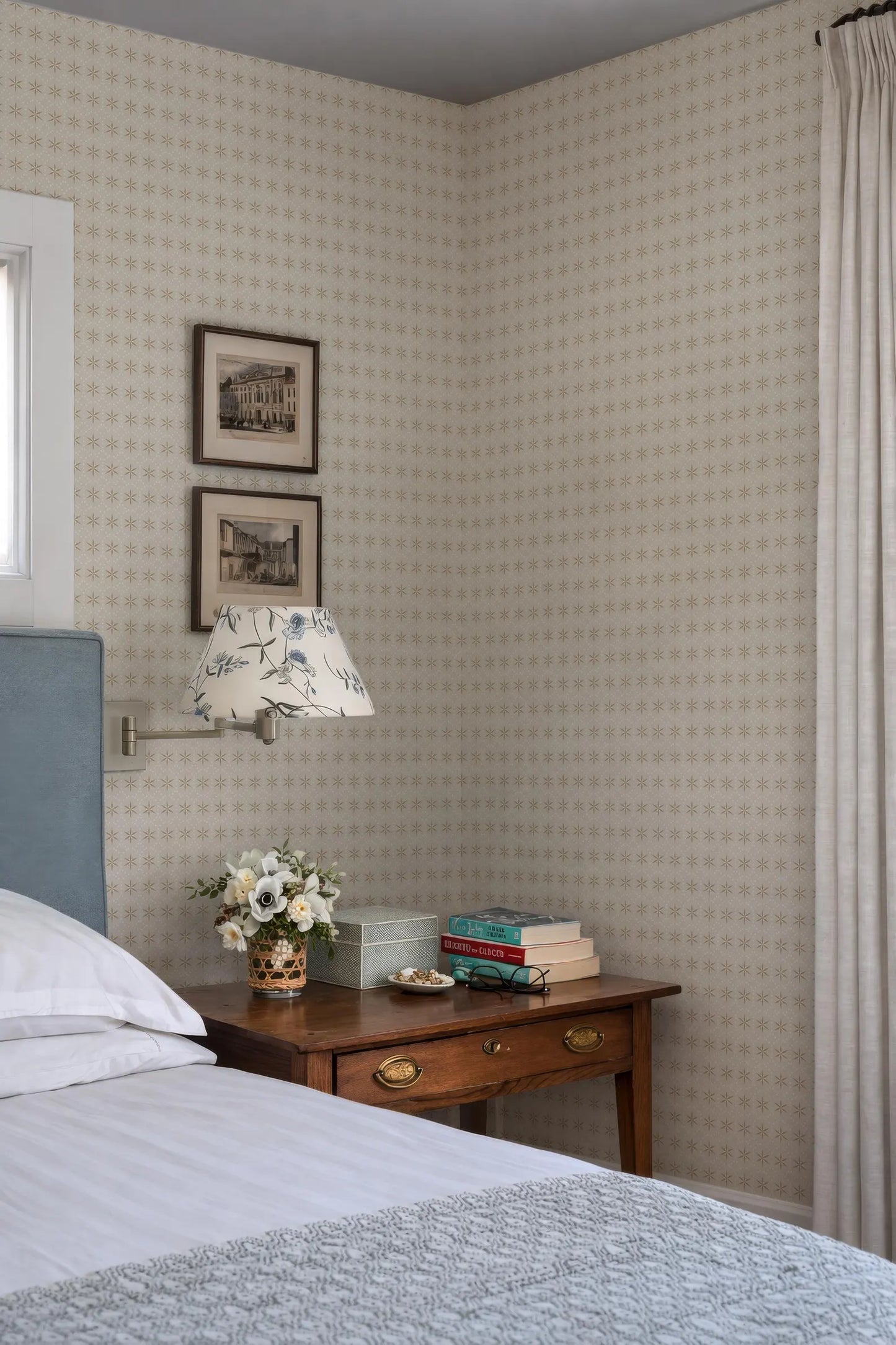 a cozy bedroom corner with a bed, a wooden nightstand, and a lamp. The wall behind the nightstand has a patterned wallpaper, and there are two framed pictures hanging on it. The nightstand also has a vase of flowers and a stack of books.
