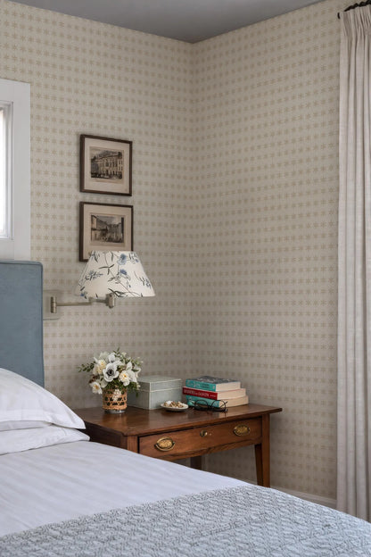 a cozy bedroom corner with a bed, a wooden nightstand, and a lamp. The wall behind the nightstand has a patterned wallpaper, and there are two framed pictures hanging on it. The nightstand also has a vase of flowers and a stack of books.