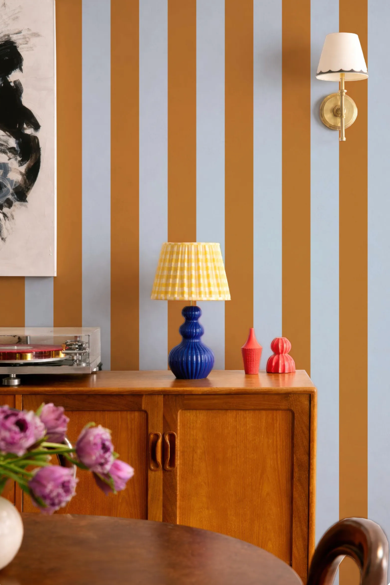 a wooden cabinet with a blue lamp and a red vase on top, set against a striped wall. The cabinet is positioned next to a table with a vase of purple flowers.