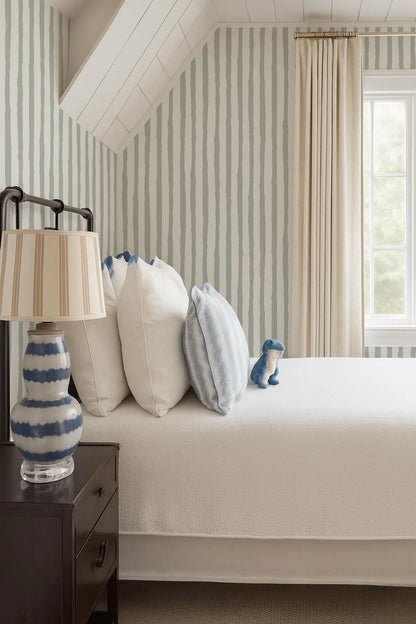 a cozy bedroom with a bed, a nightstand, and a lamp. The bed is adorned with white pillows and a blue and white striped pillowcase. The nightstand has a lamp with a white shade and a blue and white striped base. The room has a window with beige curtains, and a stuffed animal is placed on the bed.