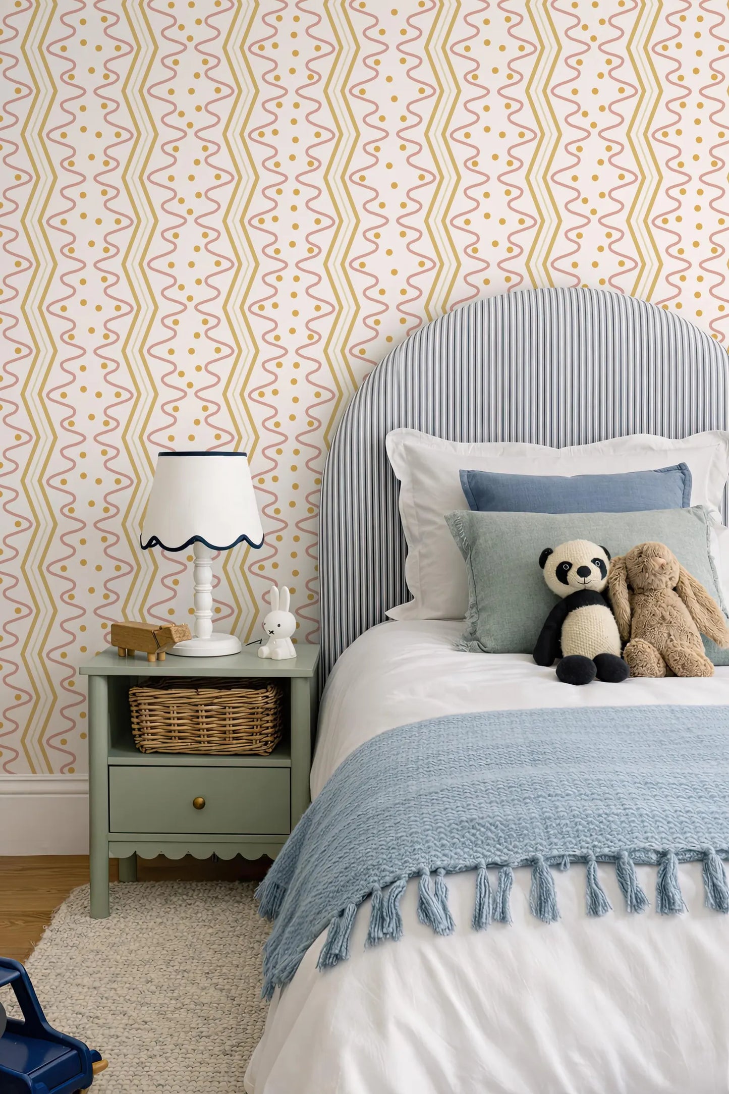 a cozy bedroom scene with a bed, nightstand, and stuffed animals. The bed has a blue and white striped headboard and a blue blanket. The nightstand has a white lamp and a small basket. There are two stuffed animals, a panda and a bunny, on the bed. The walls are covered in a patterned wallpaper with yellow and orange lines.