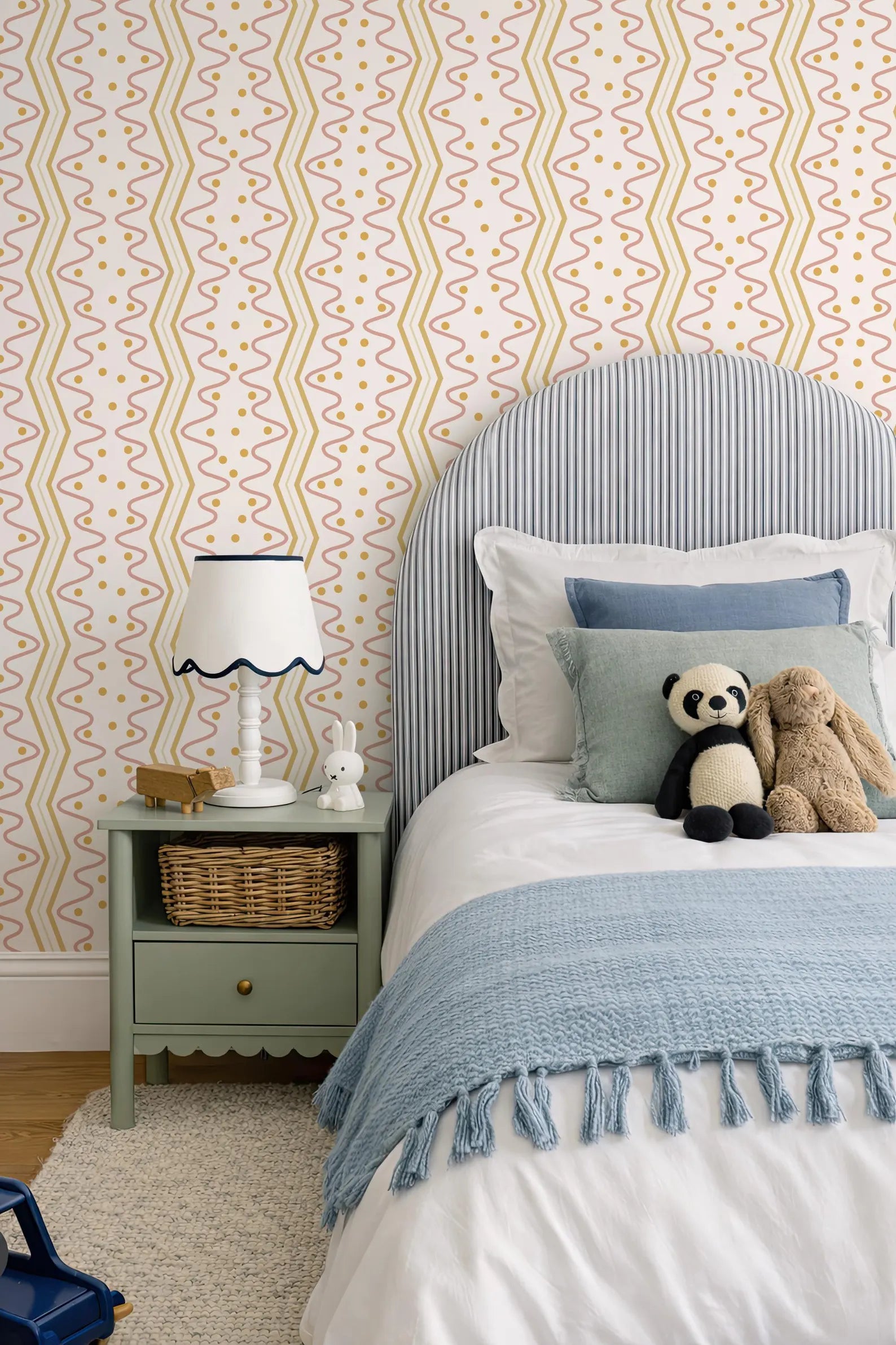 a cozy bedroom scene with a bed, nightstand, and stuffed animals. The bed has a blue and white striped headboard and a blue blanket. The nightstand has a white lamp and a small basket. There are two stuffed animals, a panda and a bunny, on the bed. The walls are covered in a patterned wallpaper with yellow and orange lines.