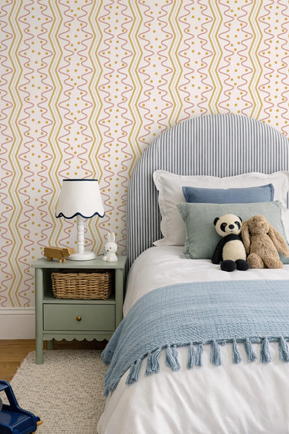 a cozy bedroom scene with a bed, nightstand, and stuffed animals. The bed has a blue and white striped headboard and a blue blanket. The nightstand has a white lamp and a small basket. There are two stuffed animals, a panda and a bunny, on the bed. The walls are covered in a patterned wallpaper with yellow and orange lines.