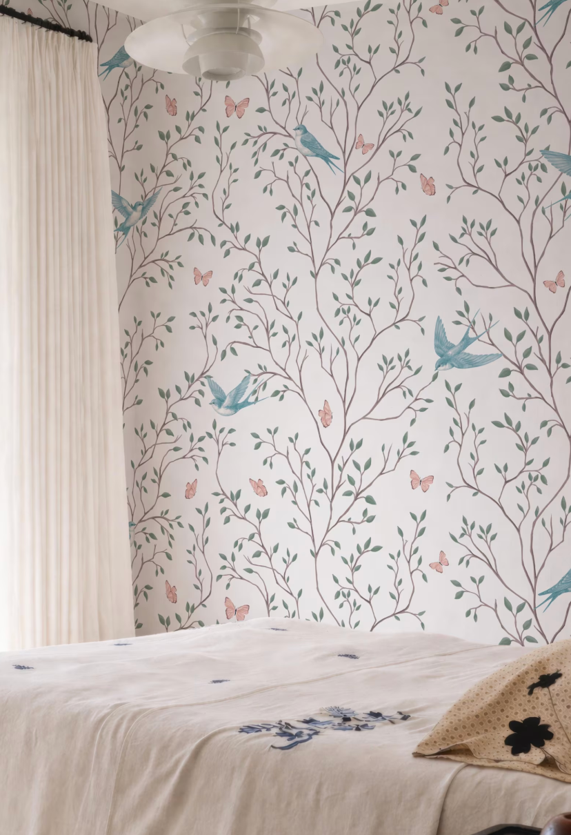 a bedroom with a white bed and a wall with a floral wallpaper featuring butterflies and leaves. The room has a window with white curtains and a ceiling fan.