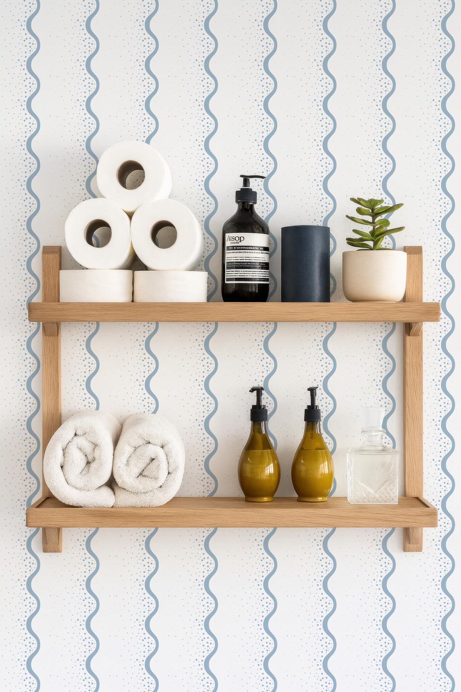 a wooden shelf with various toiletries and decorative items, including rolled towels, a soap dispenser, a bottle of lotion, and a small potted plant.