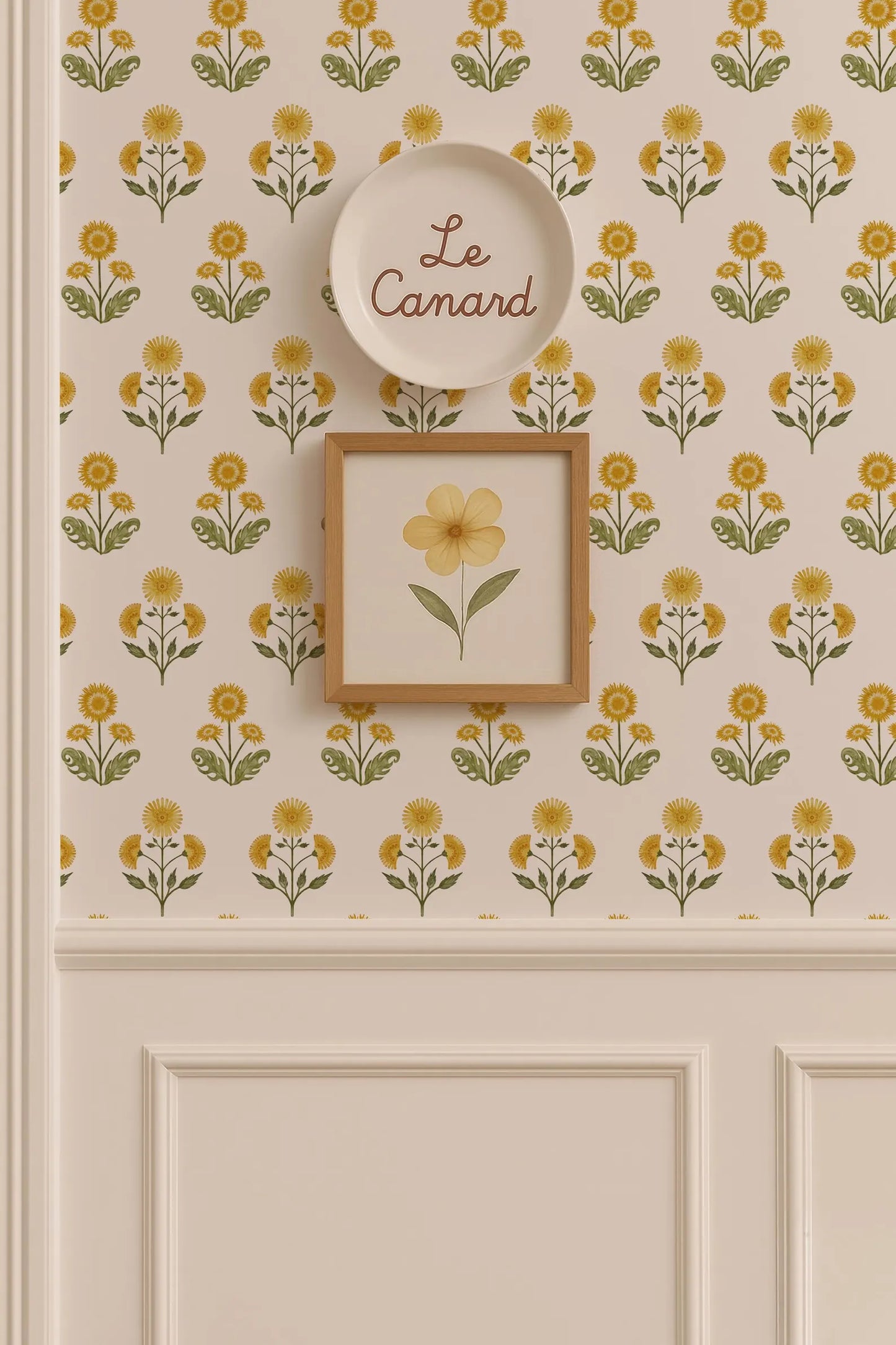 a wall with a floral pattern and a framed picture of a yellow flower. Above the wall, there is a plate with the word "Le Camand" written on it.