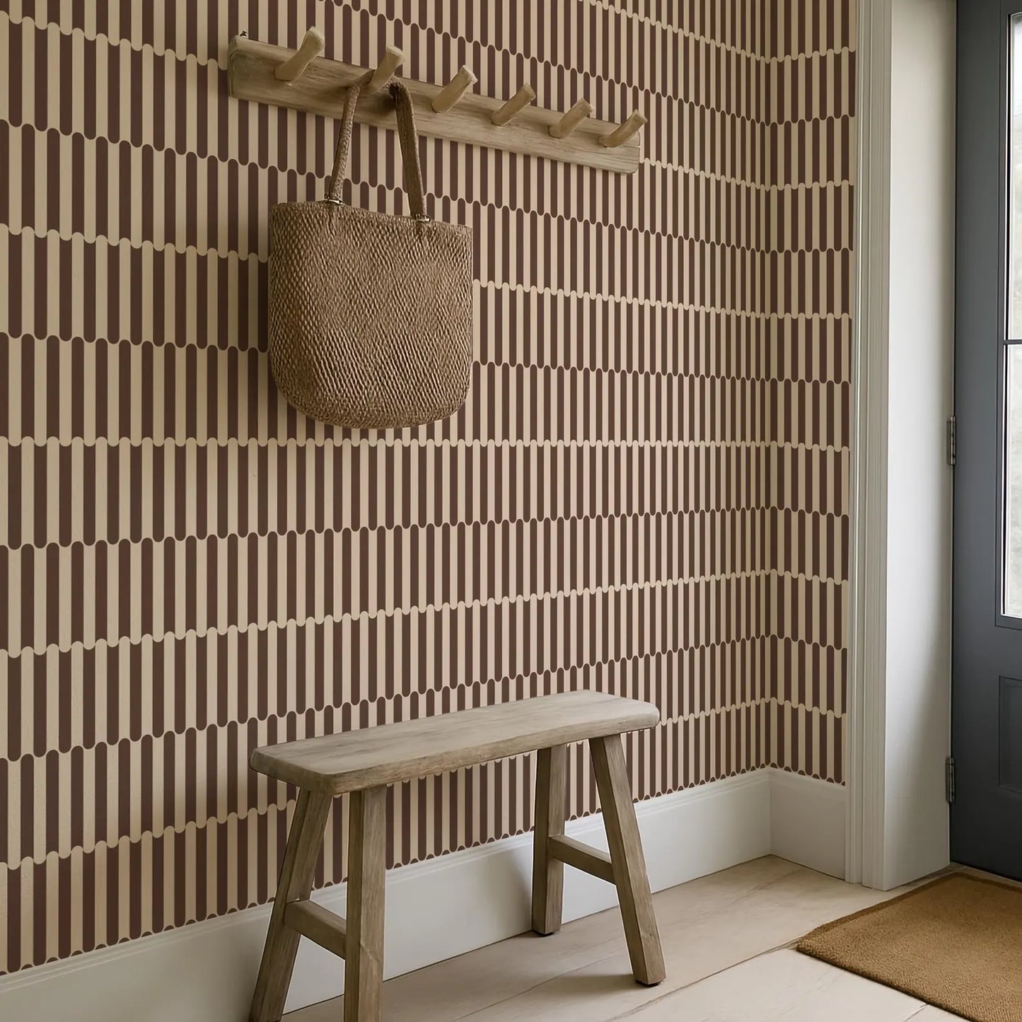 a wooden bench situated in a room with a striped wallpaper pattern. Above the bench, there is a wooden coat rack with a woven bag hanging on it. The room has a white door and a brown rug on the floor.
