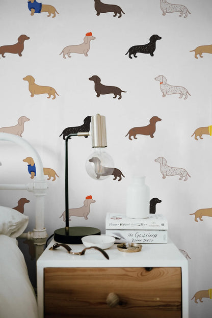a white nightstand with a lamp, a stack of books, and a pair of glasses on it. The wall behind the nightstand has a repeating pattern of dachshund illustrations in various colors and sizes.
