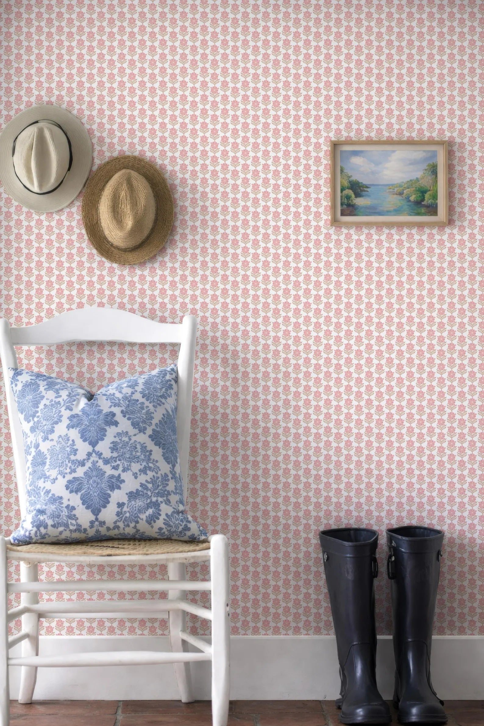 a white chair with a blue and white floral pillow, a pair of black rain boots, and a framed landscape painting on a pink and white patterned wall.