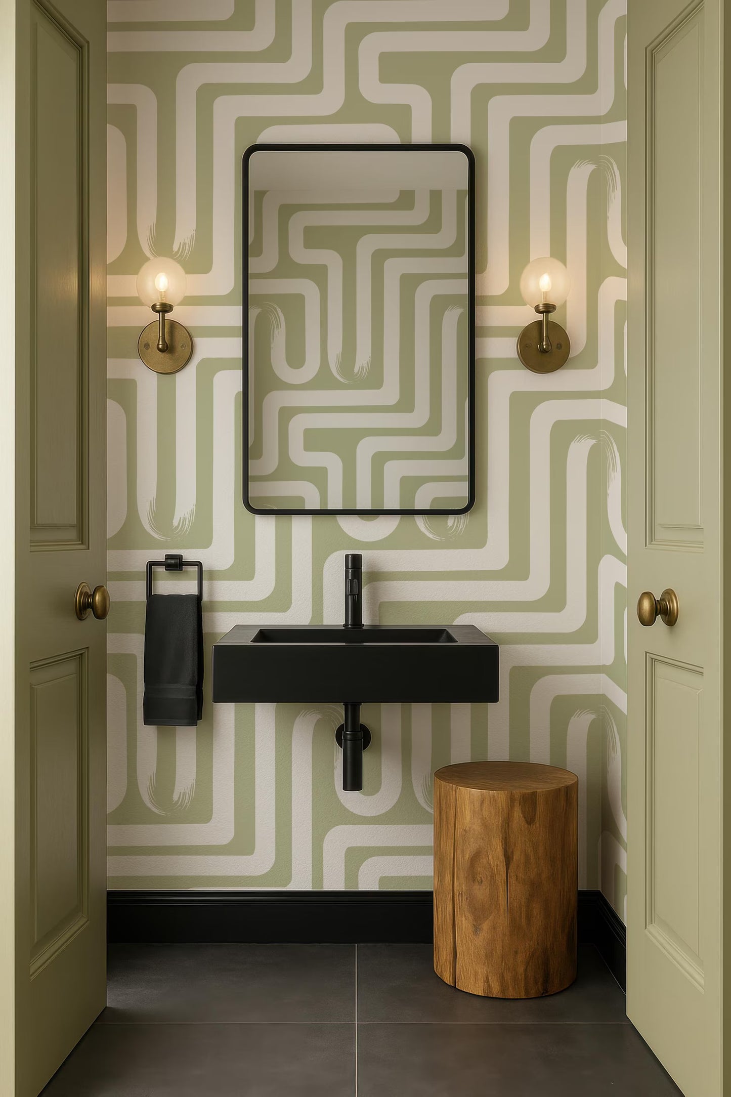 a bathroom with a black sink, a mirror, and two wall sconces. The walls are covered in a green and white patterned wallpaper, and there is a wooden stool in the corner.