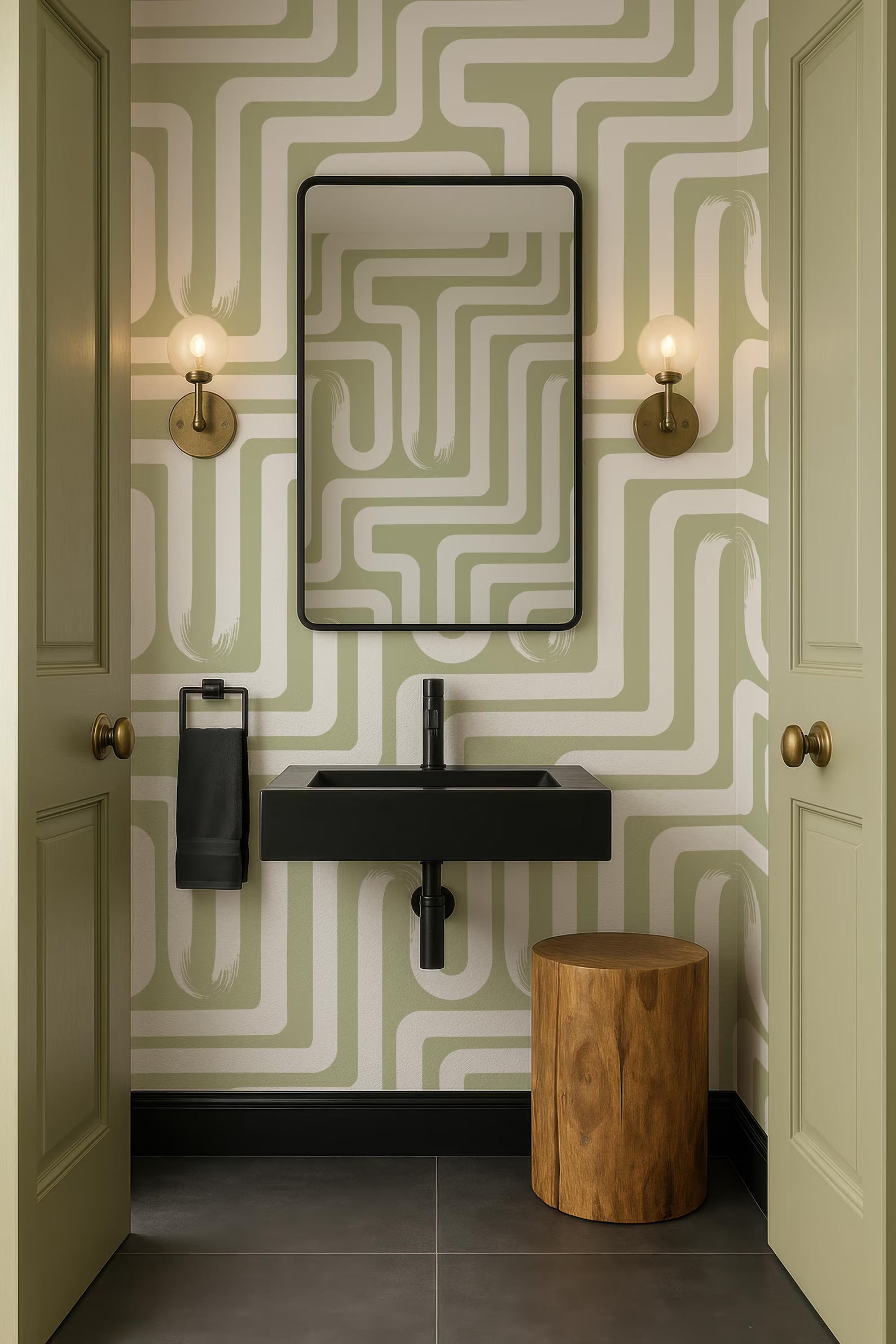 a bathroom with a black sink, a mirror, and two wall sconces. The walls are covered in a green and white patterned wallpaper, and there is a wooden stool in the corner.