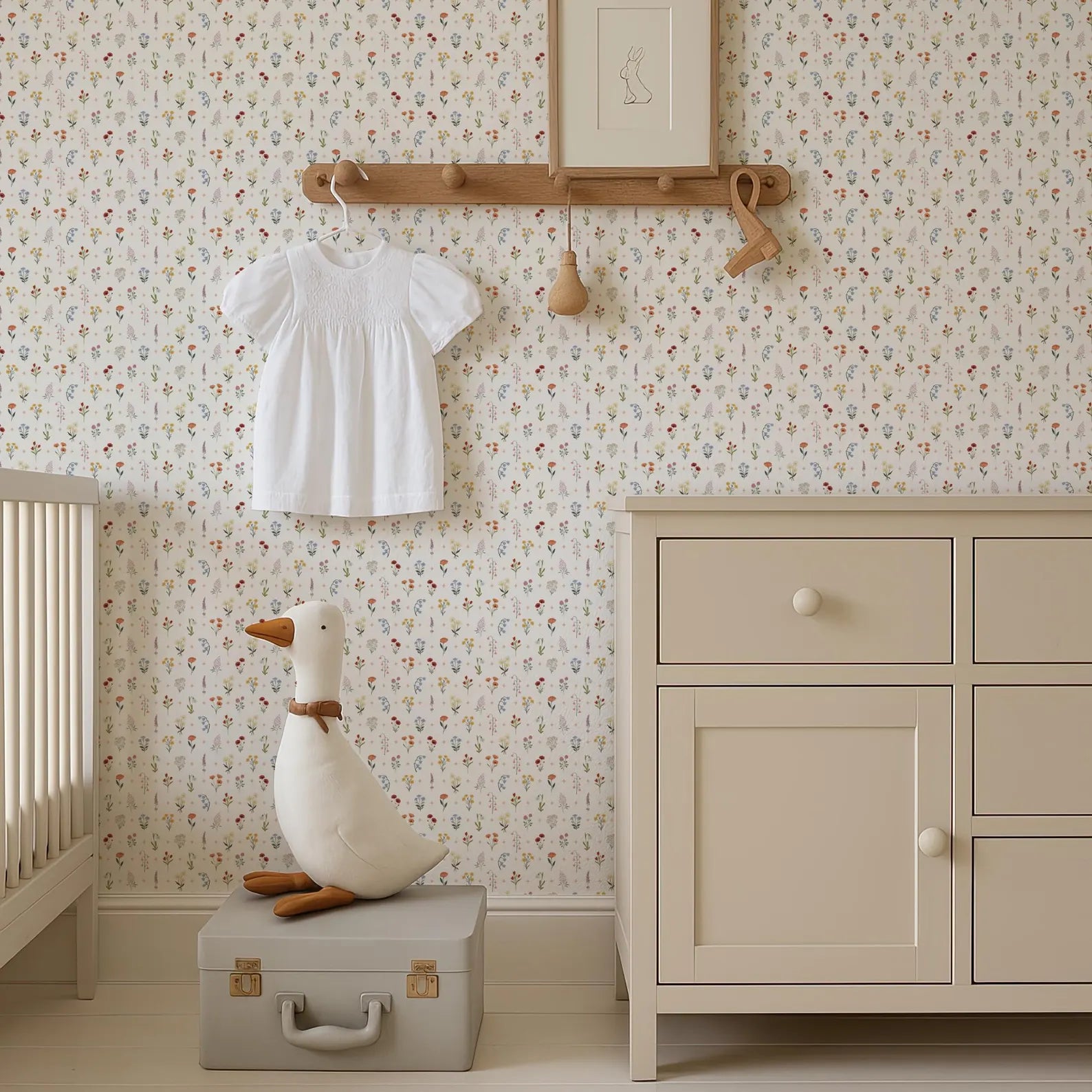 a nursery room with a white dresser, a stuffed duck toy, and a wooden hanger with a white dress hanging on it. The walls are covered in a floral wallpaper, and there is a picture frame hanging on the wall.