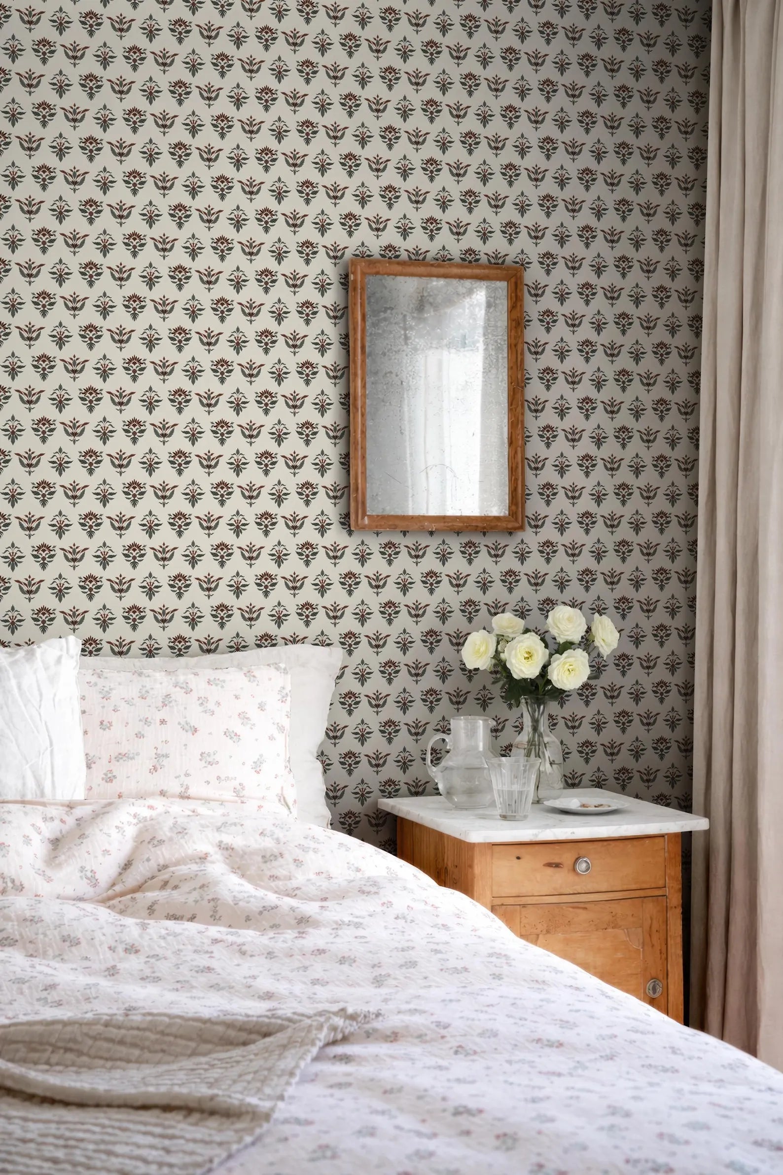 a cozy bedroom scene with a bed, a wooden nightstand, and a mirror on the wall. The bed is covered in a white and floral comforter, and there are two white pillows on it. The nightstand has a vase with white flowers, and a small picture frame is hanging above it. The mirror is positioned above the nightstand, reflecting the room's decor.
