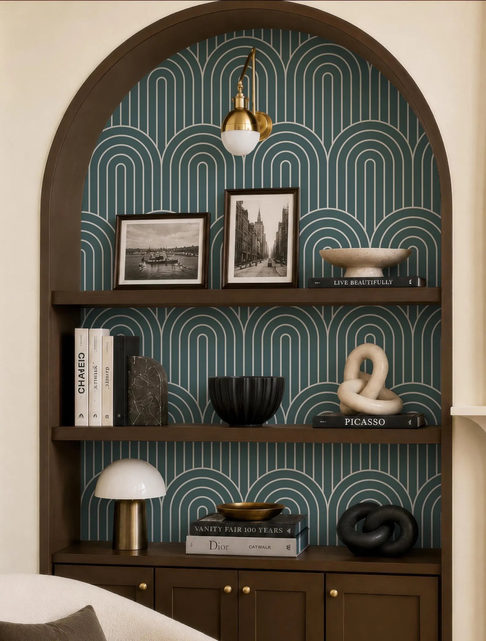 a bookshelf with a decorative archway, displaying various items such as books, a lamp, a bowl, and a sculpture. The bookshelf is set against a patterned wallpaper with a repeating wave design.