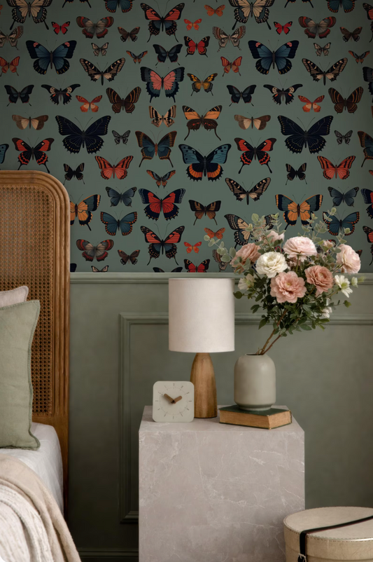 a bedroom with a wallpapered wall adorned with a pattern of colorful butterflies. The room is decorated with a wooden headboard, a lamp, a vase with flowers, and a bookshelf.