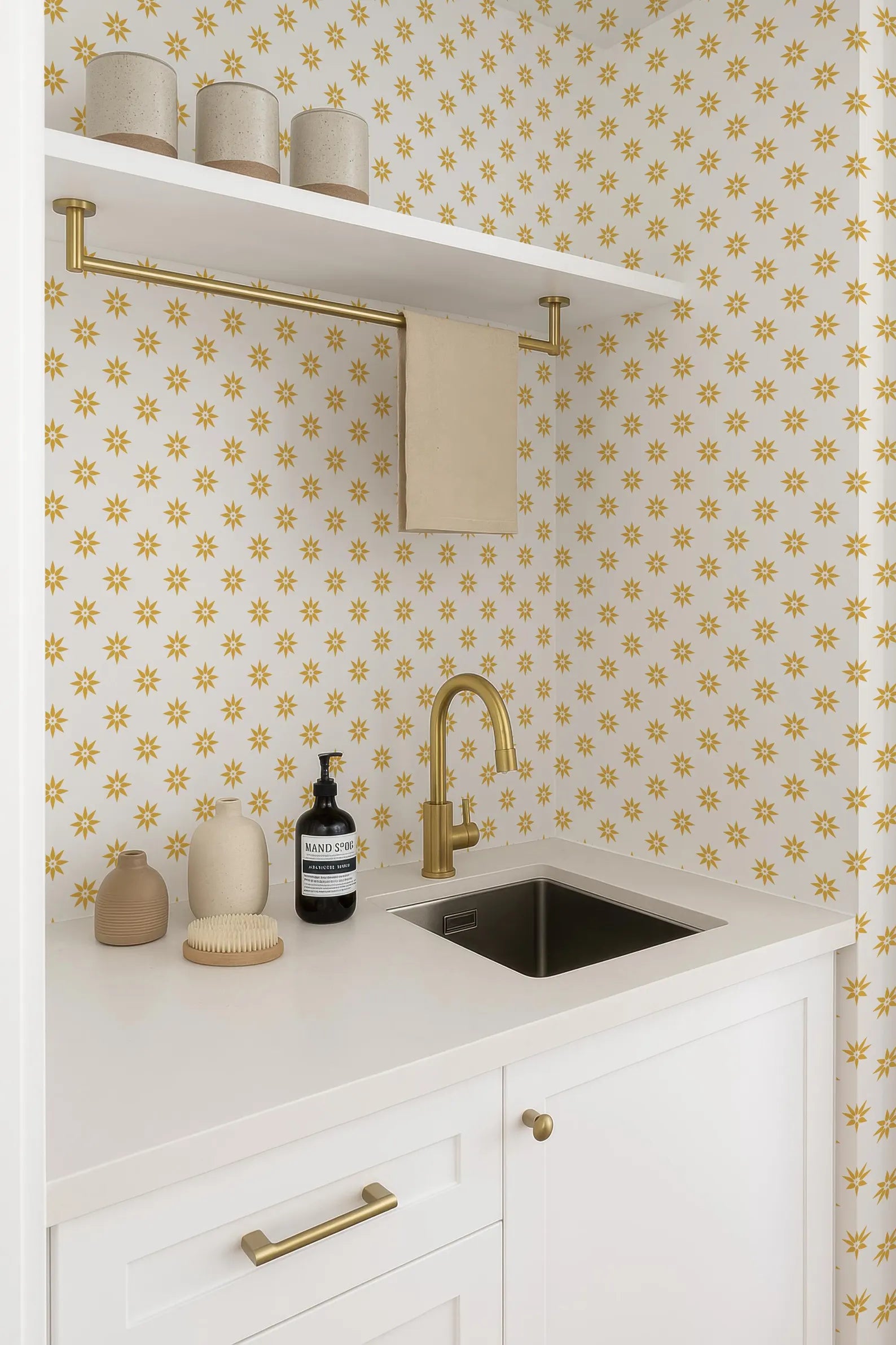 a bathroom with a white countertop, a sink, and a shelf with various items on it. The wall behind the countertop has a pattern of yellow flowers, and the overall color scheme is predominantly white and gold.