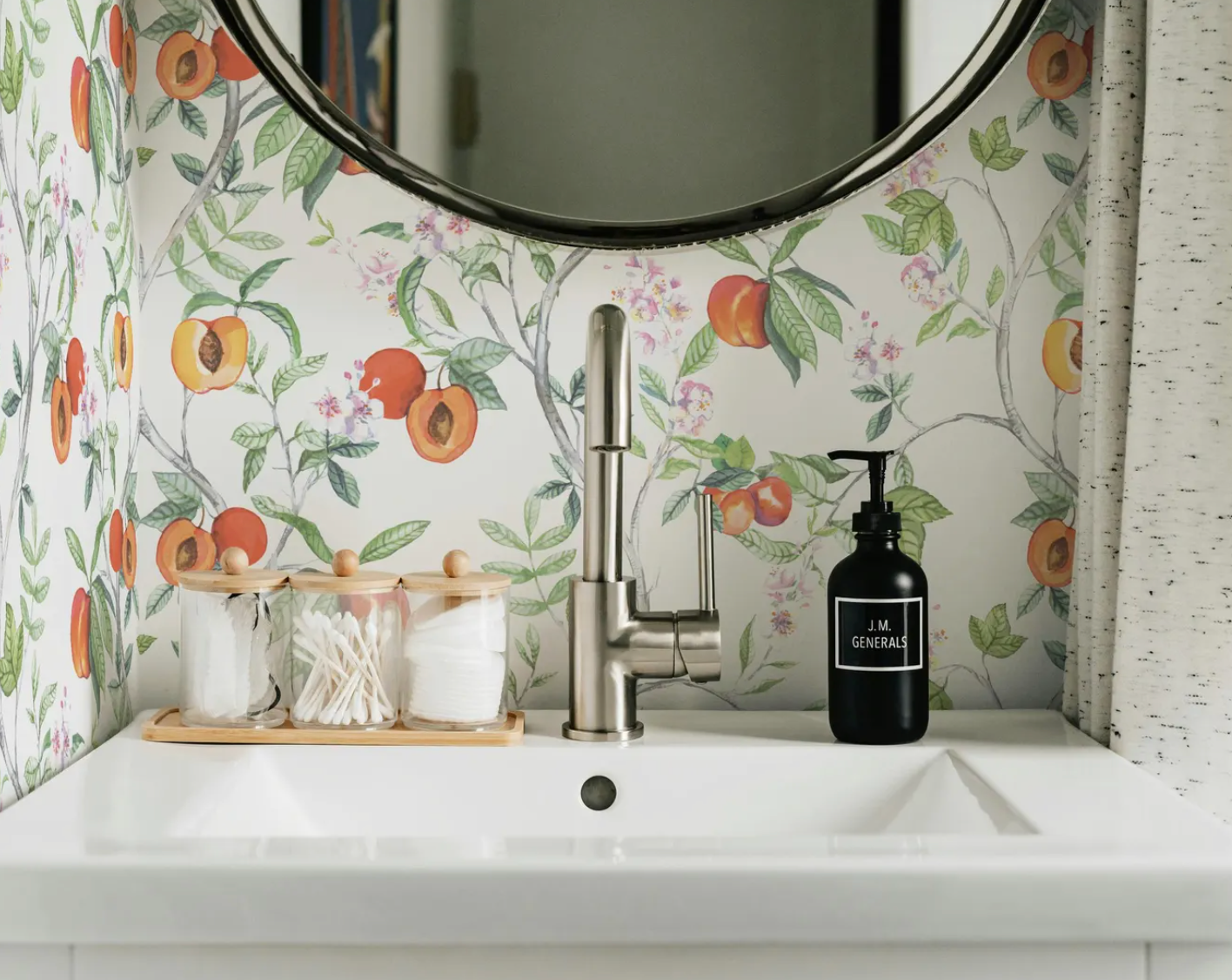 a bathroom sink with a round mirror above it, a black soap dispenser, and three small glass jars with cotton swabs. The wall behind the sink has a floral pattern, and there is a curtain on the right side of the image.