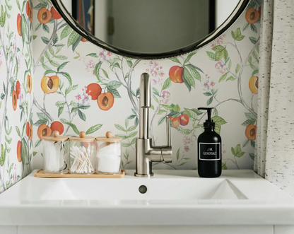 a bathroom sink with a round mirror above it, a black soap dispenser, and three small glass jars with cotton swabs. The wall behind the sink has a floral pattern, and there is a curtain on the right side of the image.