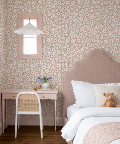 a cozy bedroom with a pink bed, a white chair, and a small table with a vase of flowers. The walls are covered in a floral wallpaper, and there is a window with a white shade. A teddy bear is resting on the bed, adding a touch of warmth and comfort to the room.