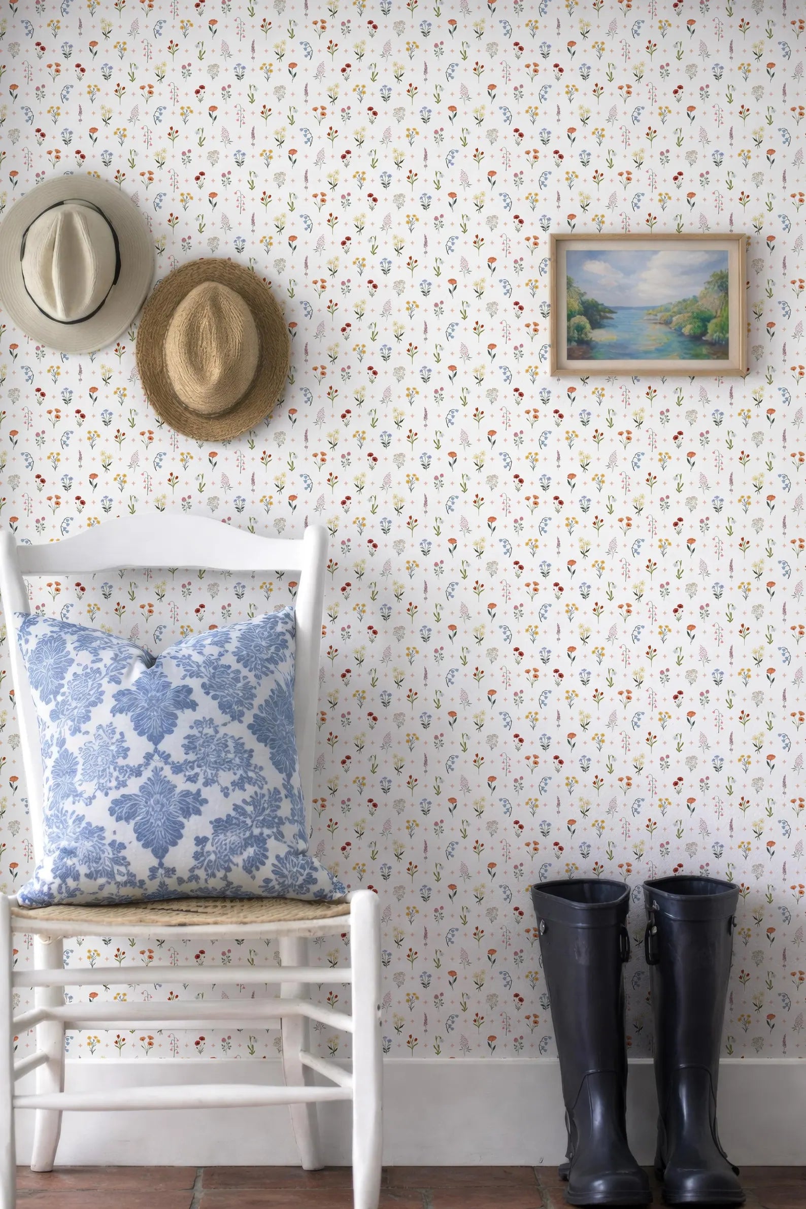 a white chair with a blue and white floral pillow, a pair of black rain boots, and a framed landscape painting on the wall.