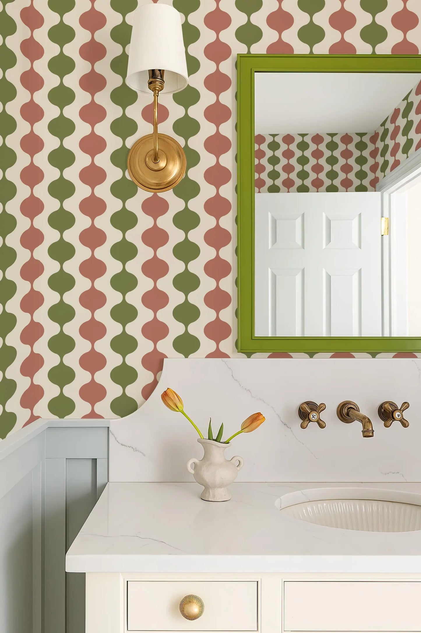 a bathroom with a white sink, a mirror, and a vase of tulips on the countertop. The walls are covered in a patterned wallpaper with a mix of green, red, and white colors. A gold-framed mirror is mounted above the sink, and a light fixture is installed above the mirror.