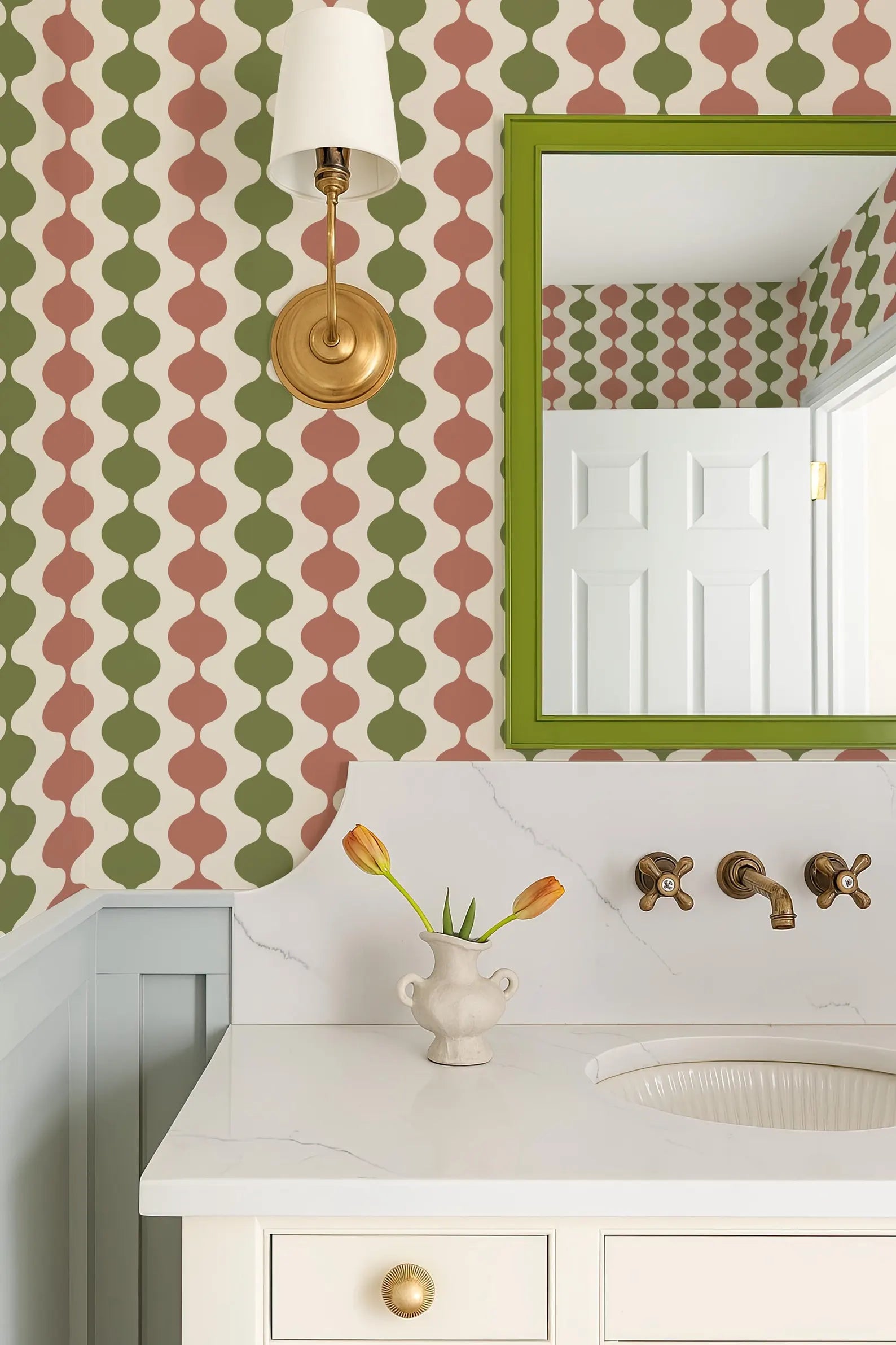 a bathroom with a white sink, a mirror, and a vase of tulips on the countertop. The walls are covered in a patterned wallpaper with a mix of green, red, and white colors. A gold-framed mirror is mounted above the sink, and a light fixture is installed above the mirror.